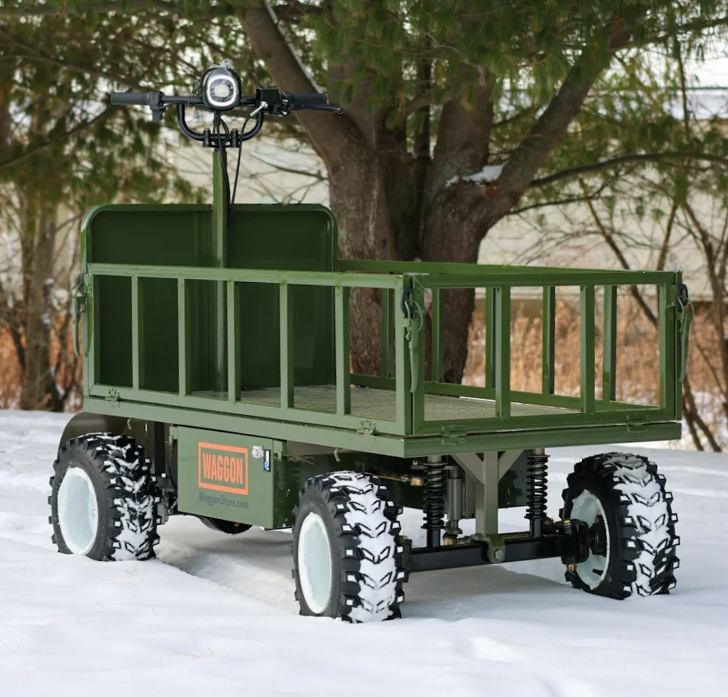 A green outdoor utility wagon with large off-road tires, parked in snow with a tree in the background.