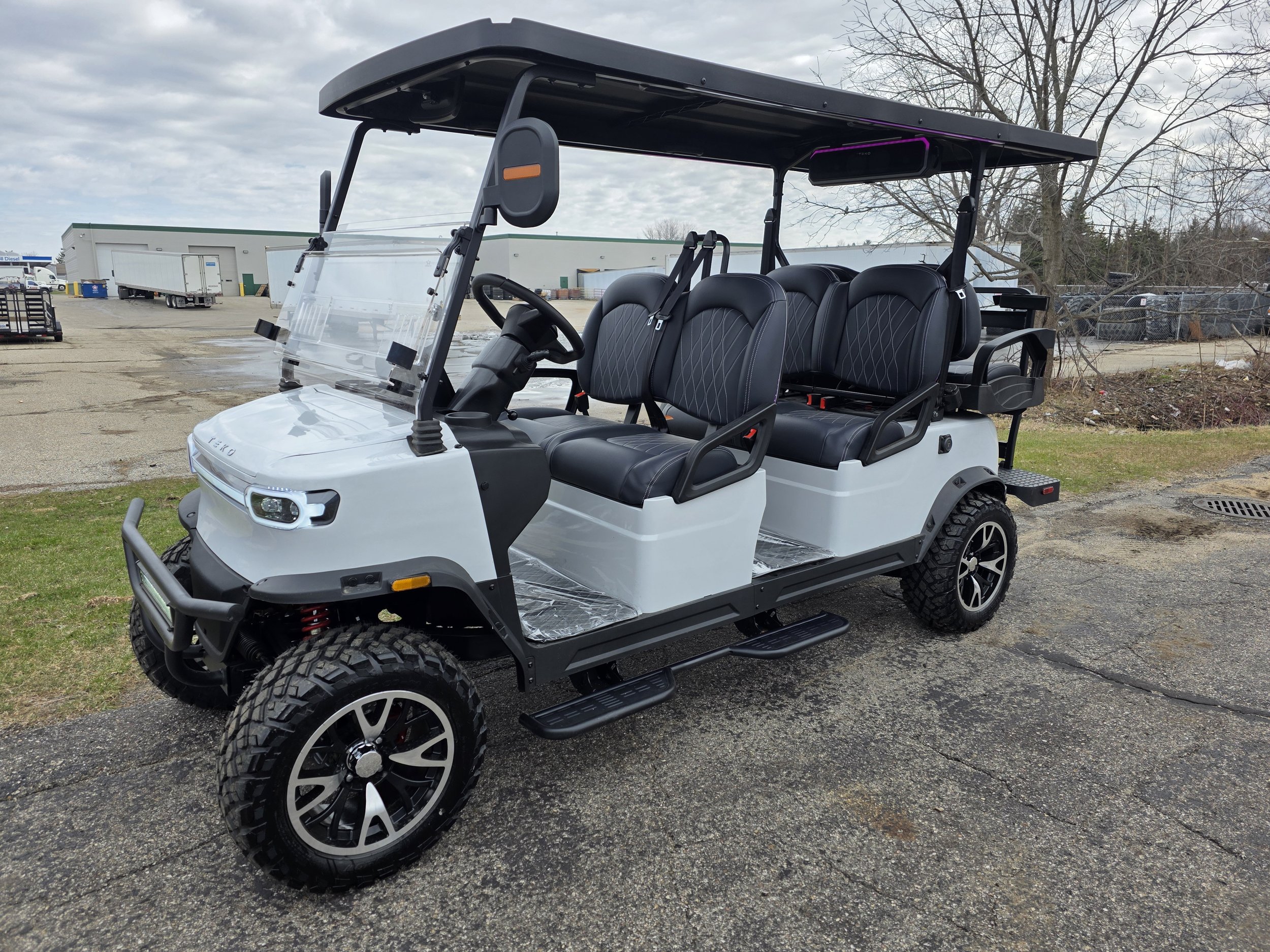 Off-road electric golf cart with four rows of black leather seats, a clear windshield, and a purple LED light on the roof, parked on a pavement.