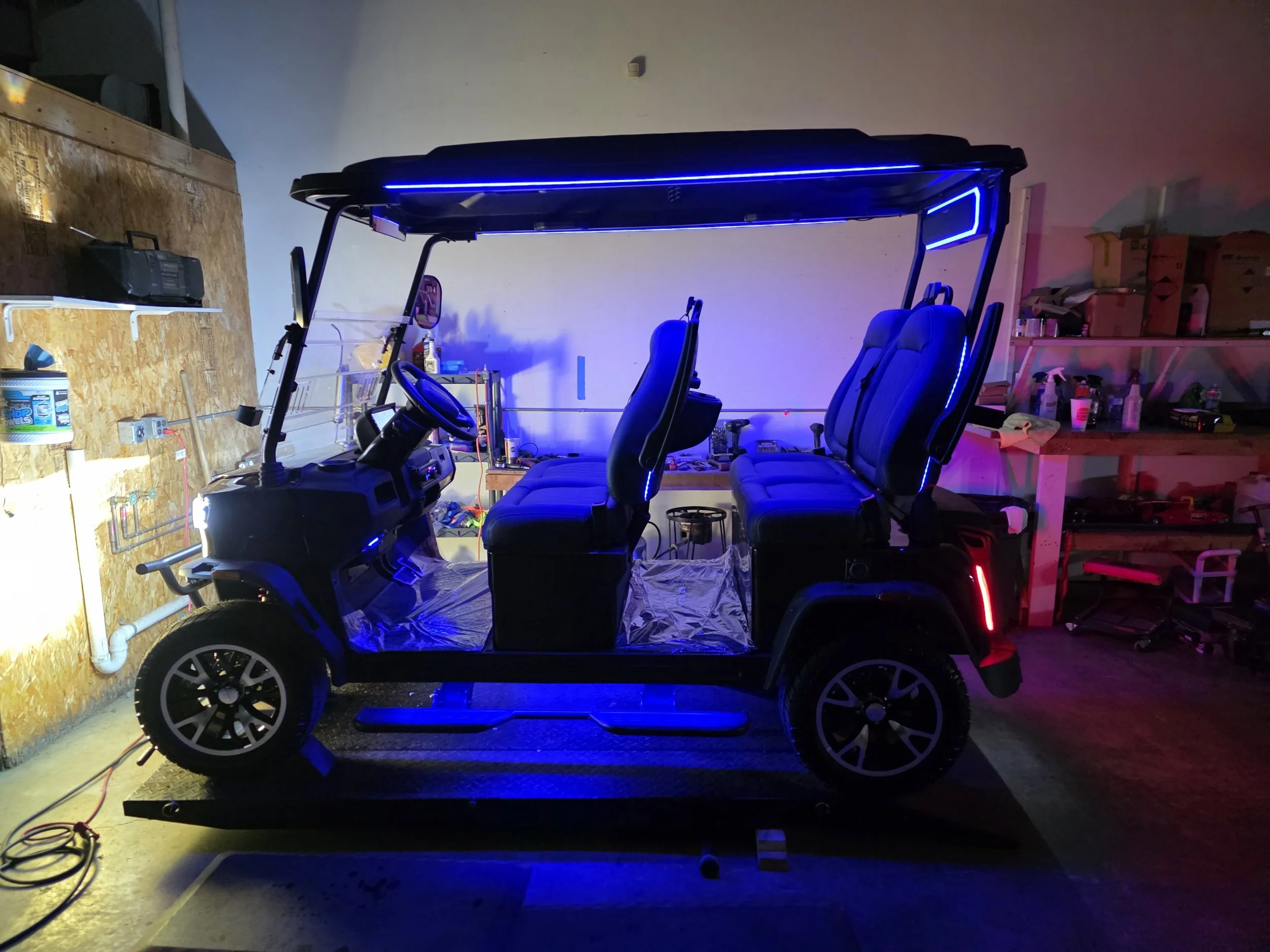 A custom electric golf cart with four rows of seats, illuminated with blue and red LED lights, parked inside a garage with shelves containing various tools and supplies.