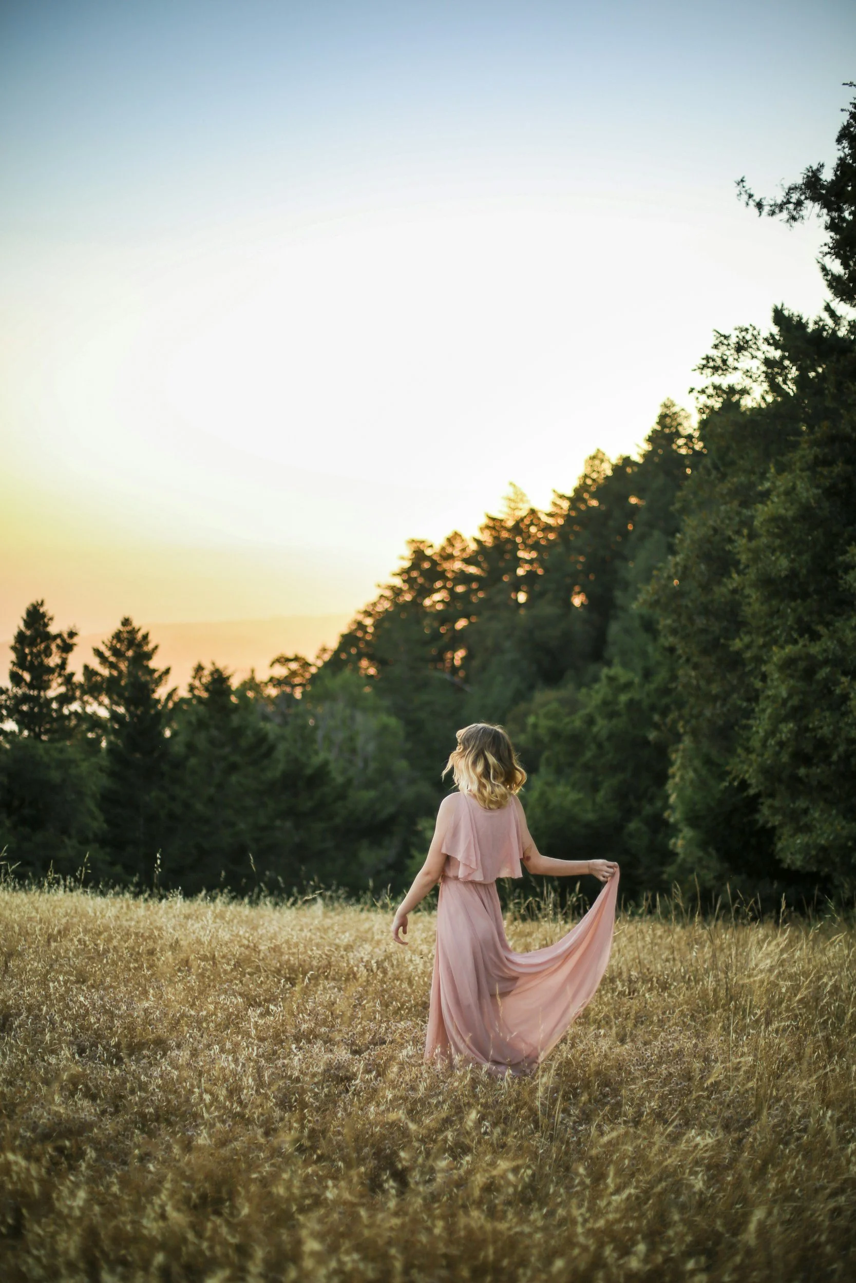 A woman in a pink dress walking through a field of tall grass at sunset, with trees in the background.