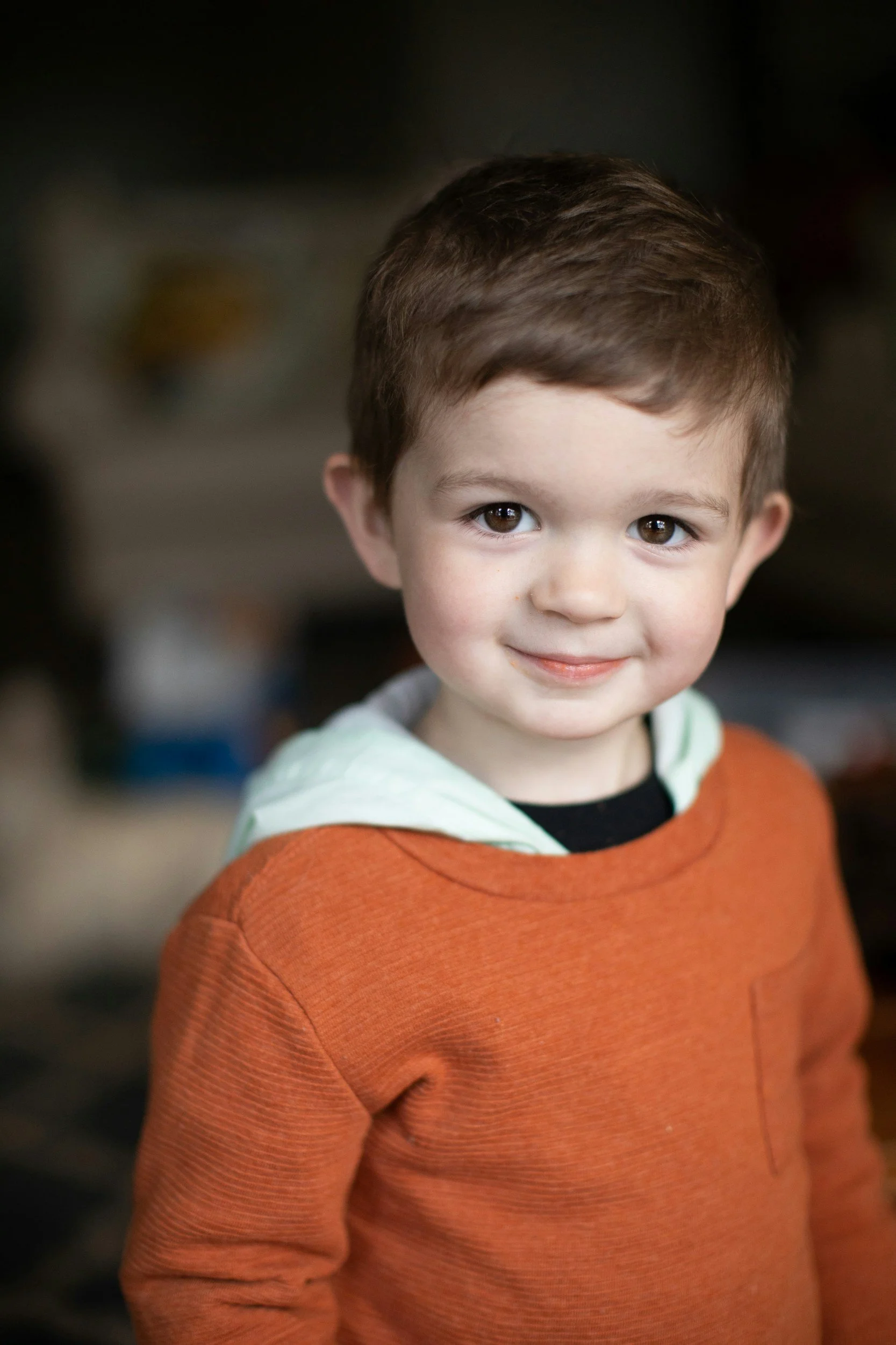 A young boy with brown hair and brown eyes, smiling at the camera, wearing an orange sweater and a black shirt underneath, with a light-colored hoodie visible under the sweater.