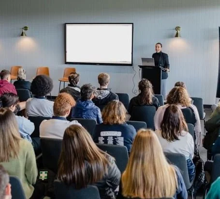 A man giving a presentation at a conference with an audience of diverse people seated and watching.