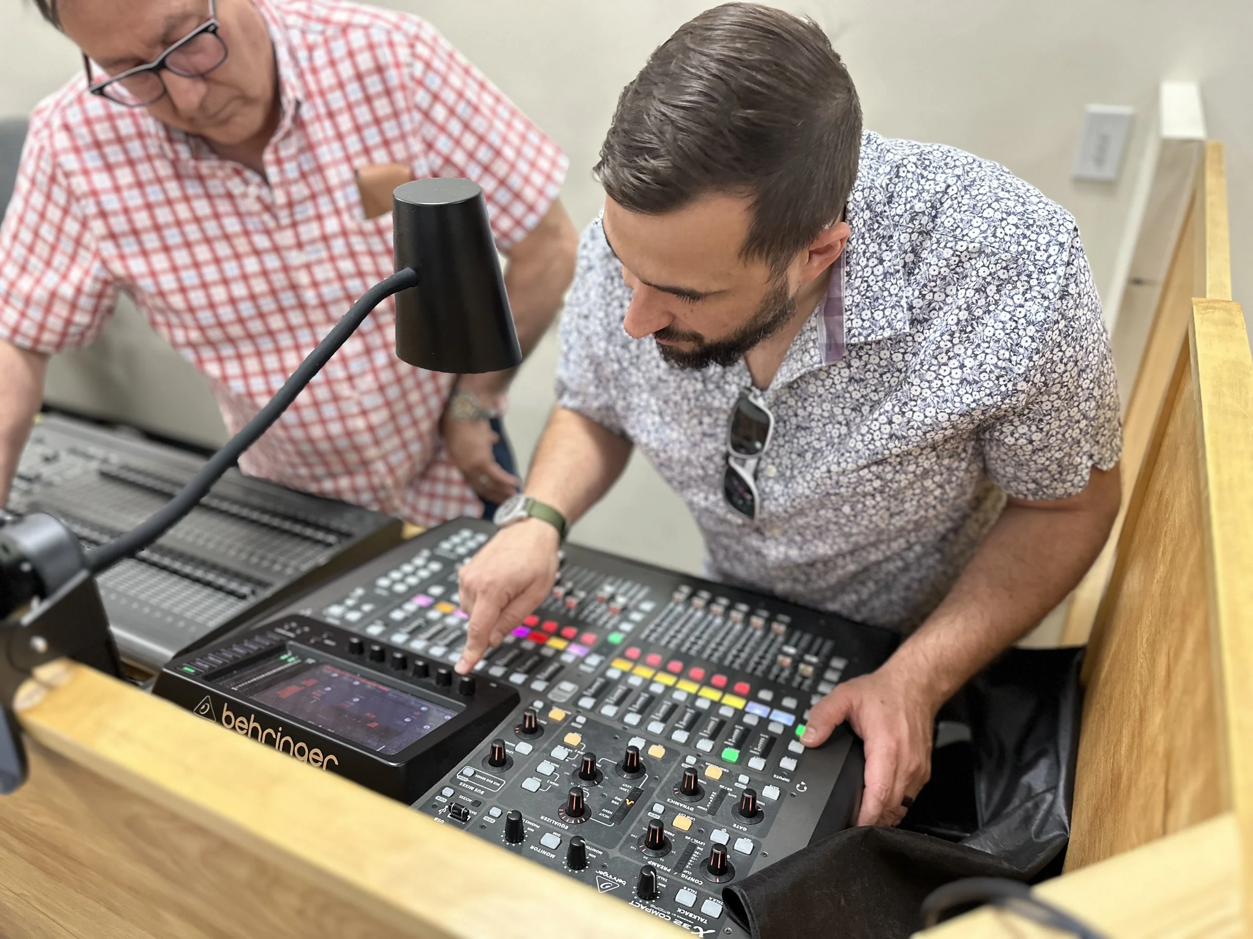Two men are working with a Behringer audio mixer, one of them points at the controls while the other looks on, in an indoor setting.