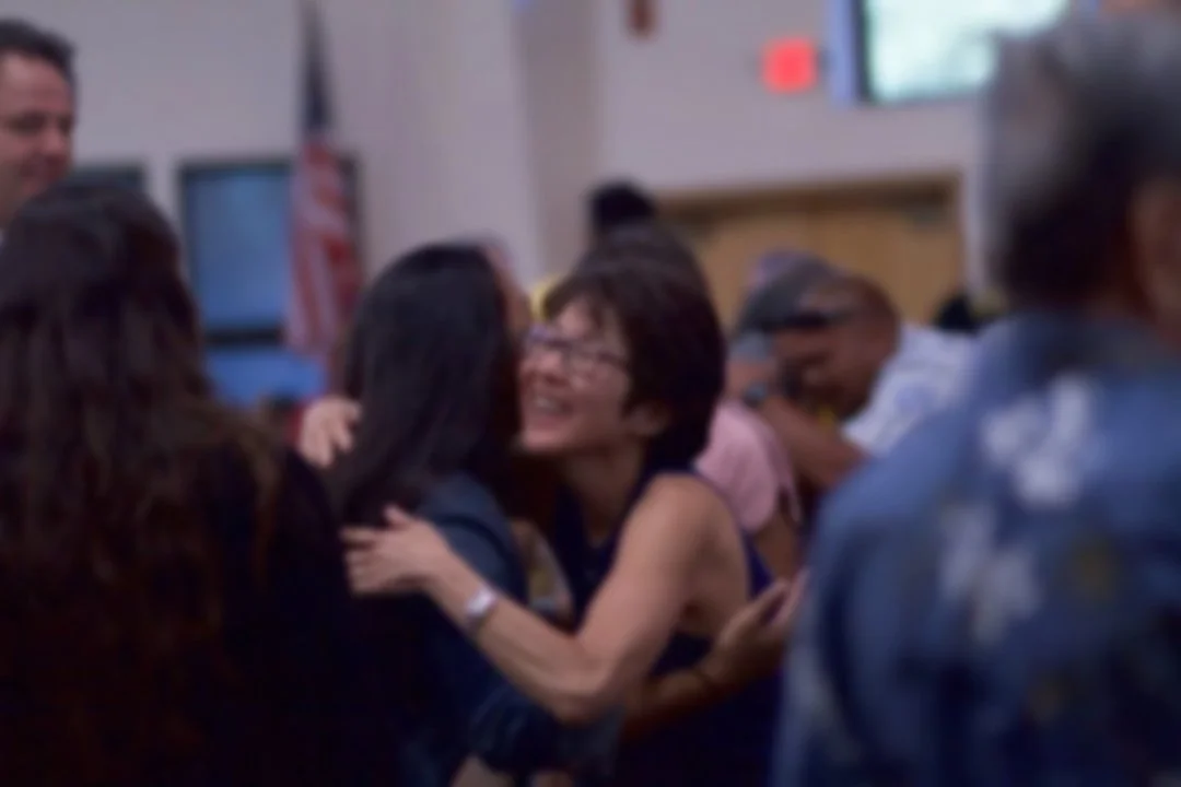 People hugging and smiling in a lively indoor setting, possibly at a social gathering or church.