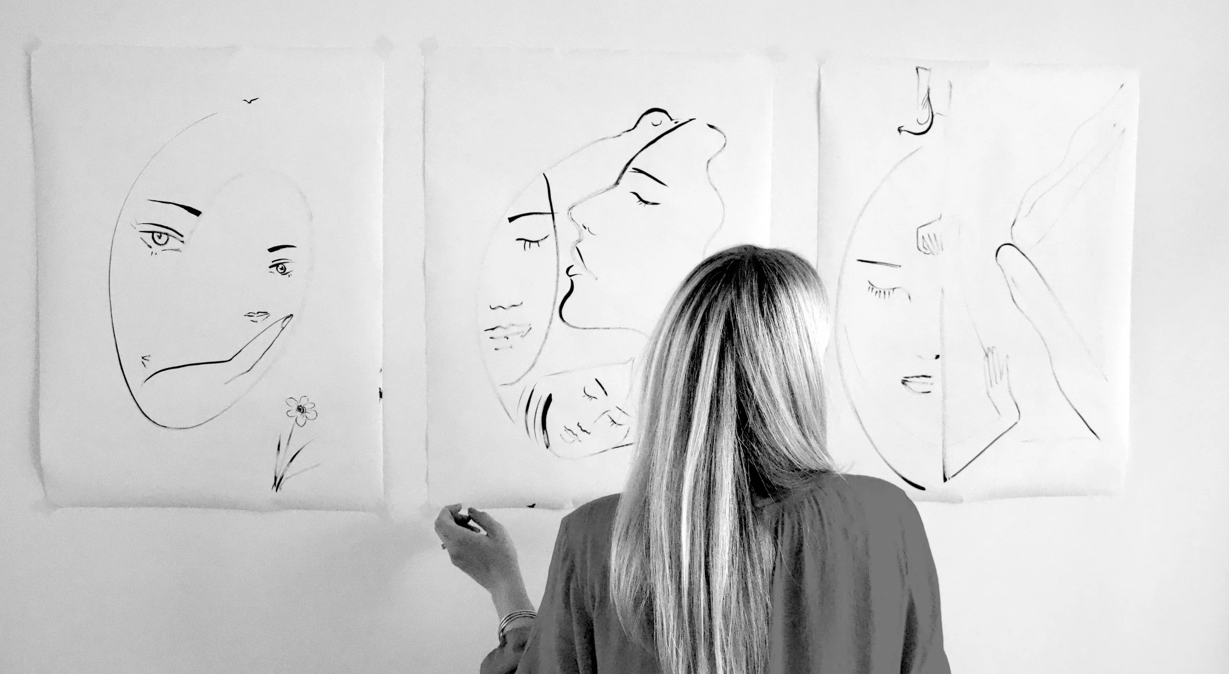 Woman observing minimalistic line drawings of faces on three pieces of paper taped to a wall.