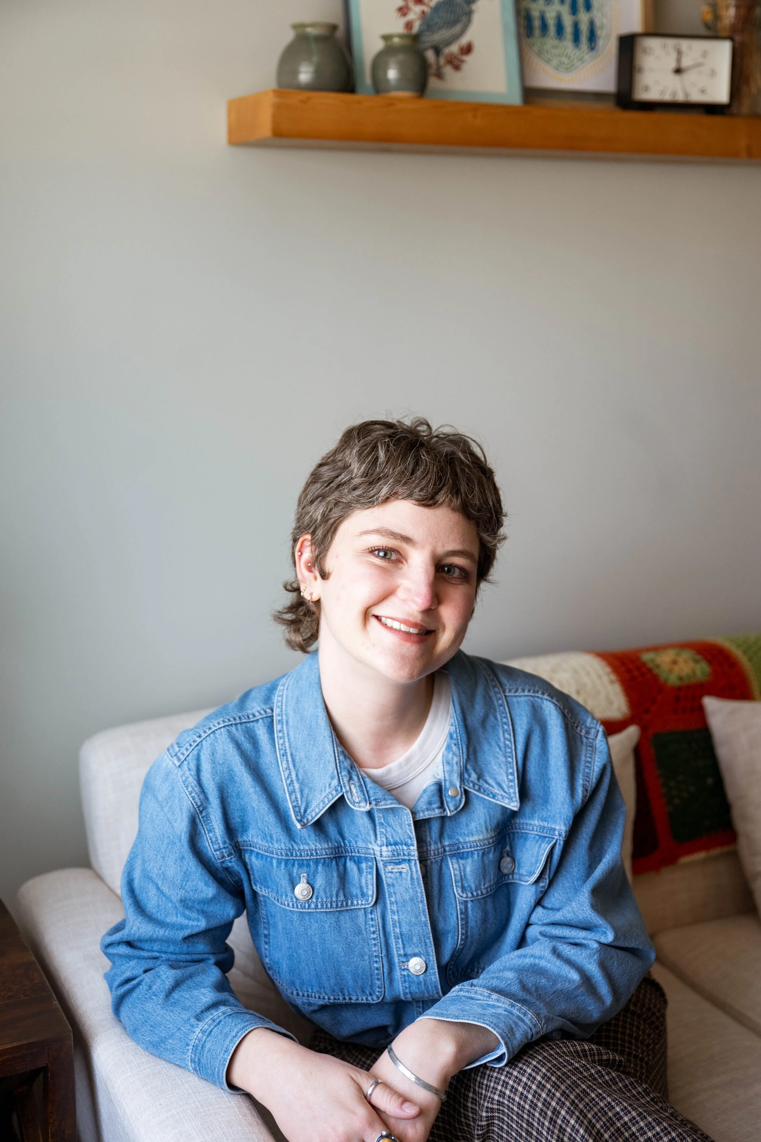 A young person with short, curly hair and light skin sitting on a white couch, smiling and wearing a blue denim jacket, in a living room with shelves, artwork, and a clock in the background.