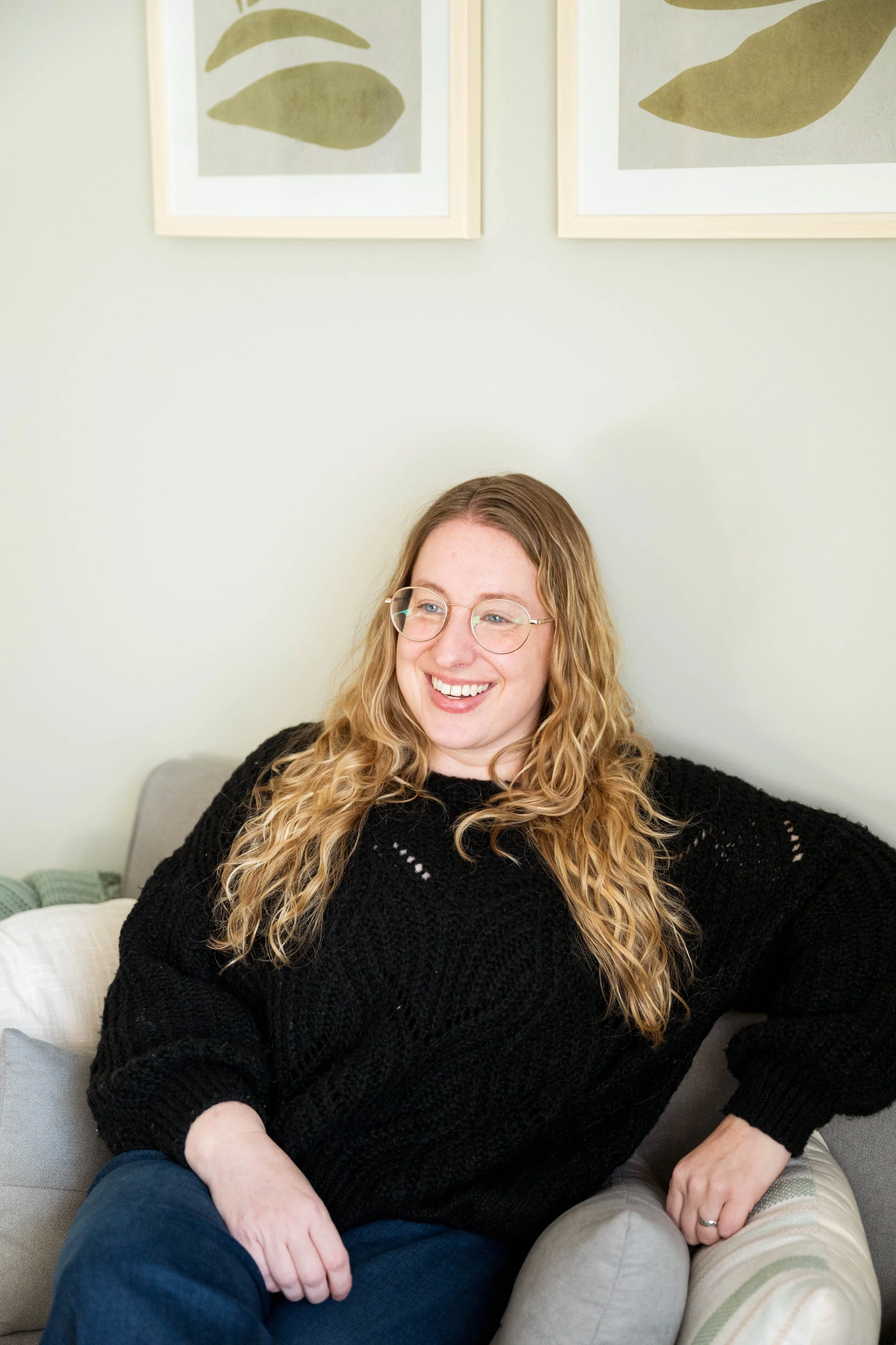 A woman with long curly blonde hair, glasses, and a black sweater, smiling while sitting on a light-colored couch in a room with pale green walls and framed artwork of leaves in the background.