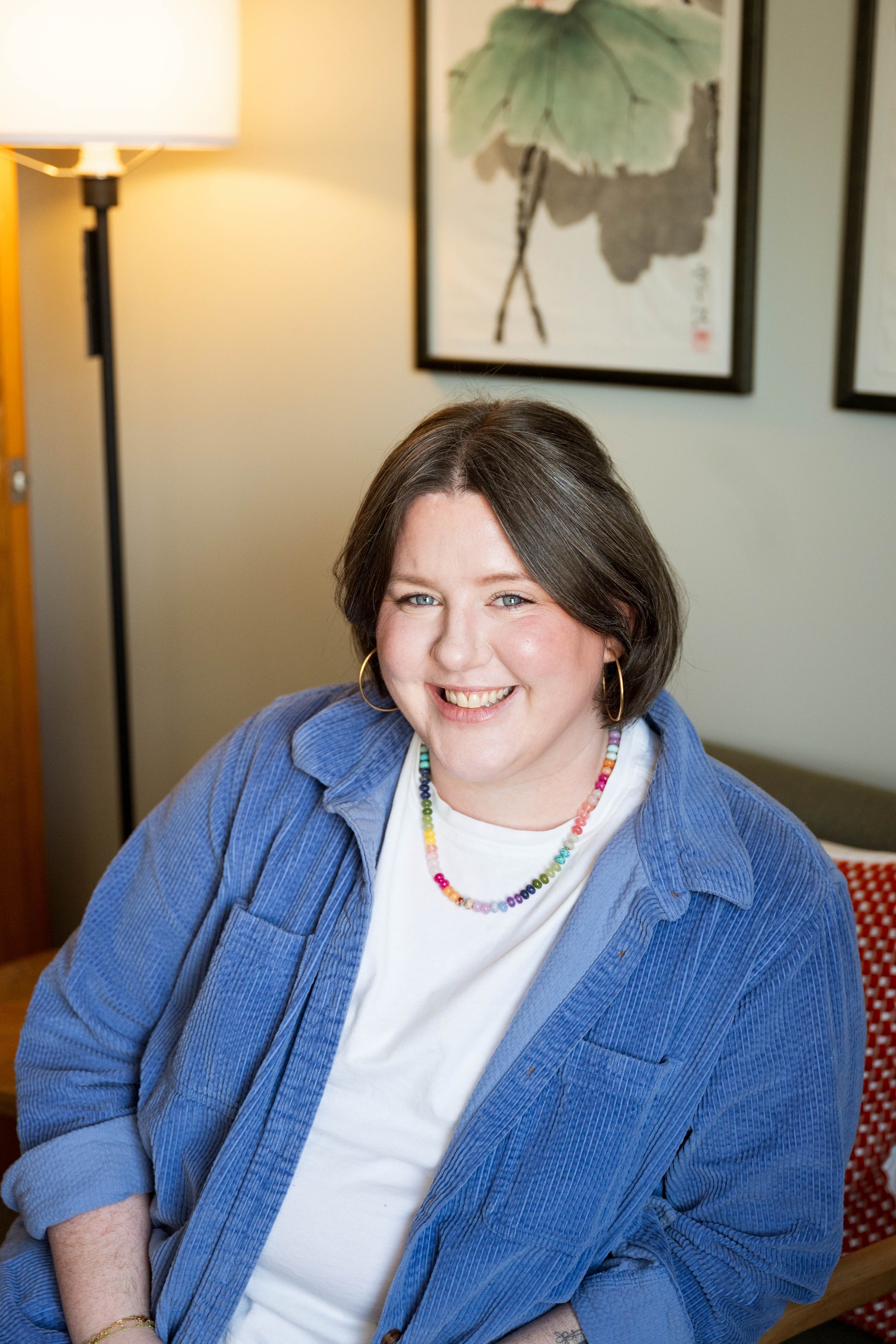 A woman with short brown hair and blue eyes sitting indoors, smiling, wearing a blue corduroy jacket, a white shirt, a colorful beaded necklace, and gold hoop earrings. There is a lamp and framed artwork of leaves on the wall behind her.