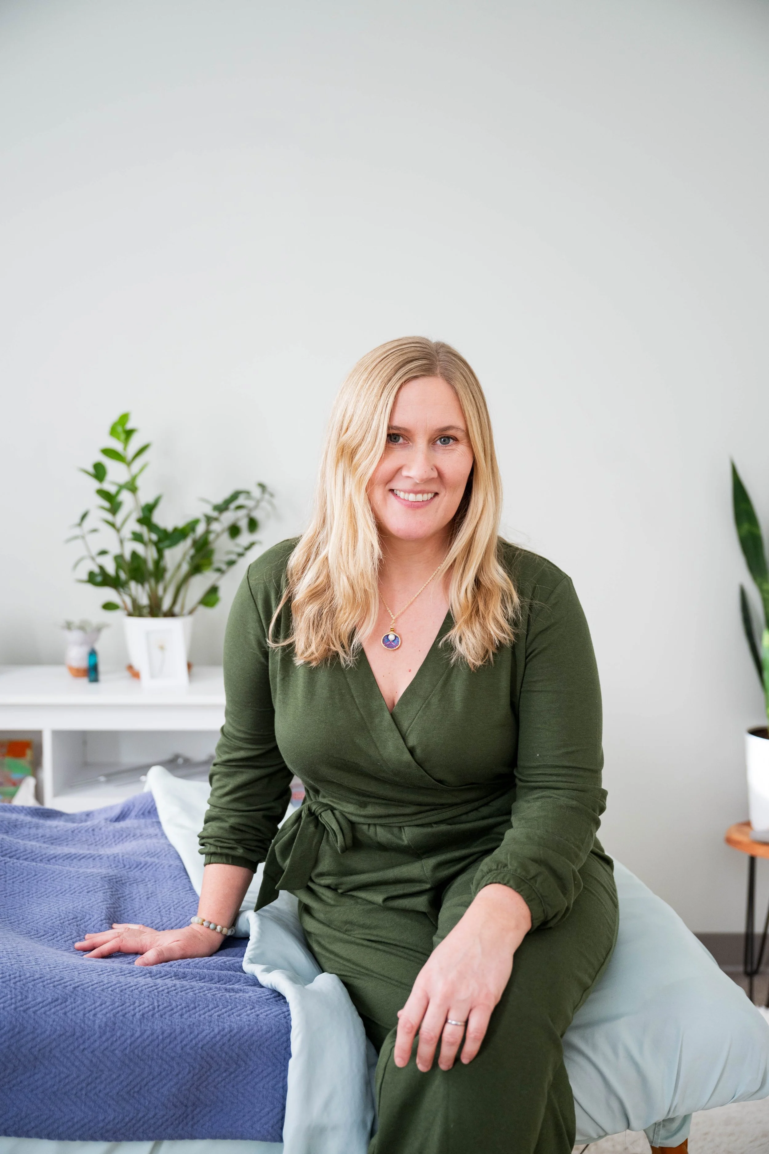 A woman with blonde hair sitting on a white textured bench, smiling, wearing a beige sweater and blue jeans, with a green plant in the background.