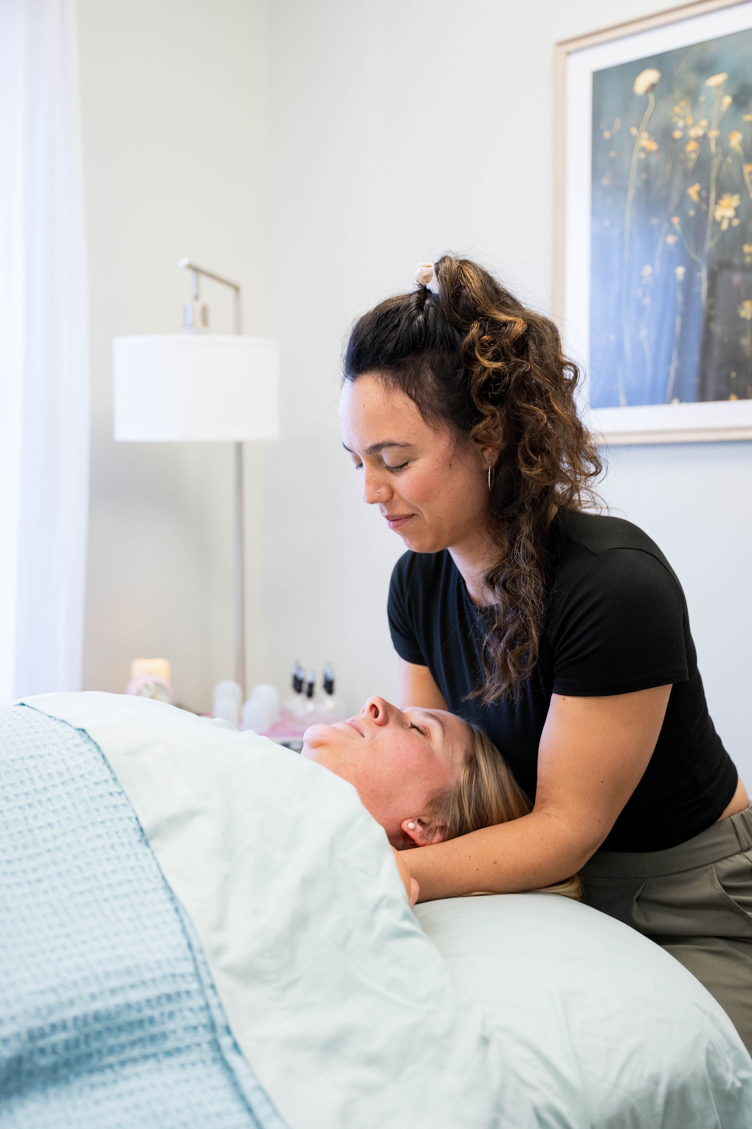 A young woman with dark hair in a braid, wearing a sleeveless pastel green dress, smiling while sitting on a yellow cushion. She has colorful beaded bracelets on her wrist and earrings, with a leafy, patterned background and a potted plant nearby.