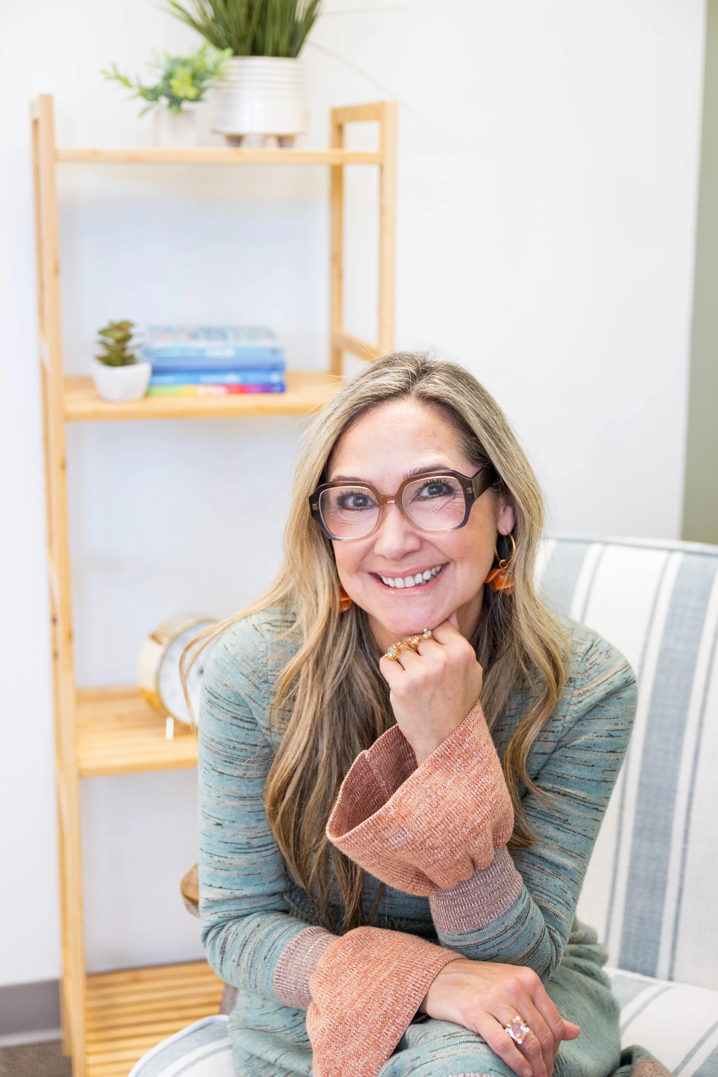 A woman with long blonde hair, glasses, and orange earrings sits on a chair, smiling at the camera with her chin resting on her hand, in a room with a striped upholstered chair and a wooden shelf with plants and books behind her.