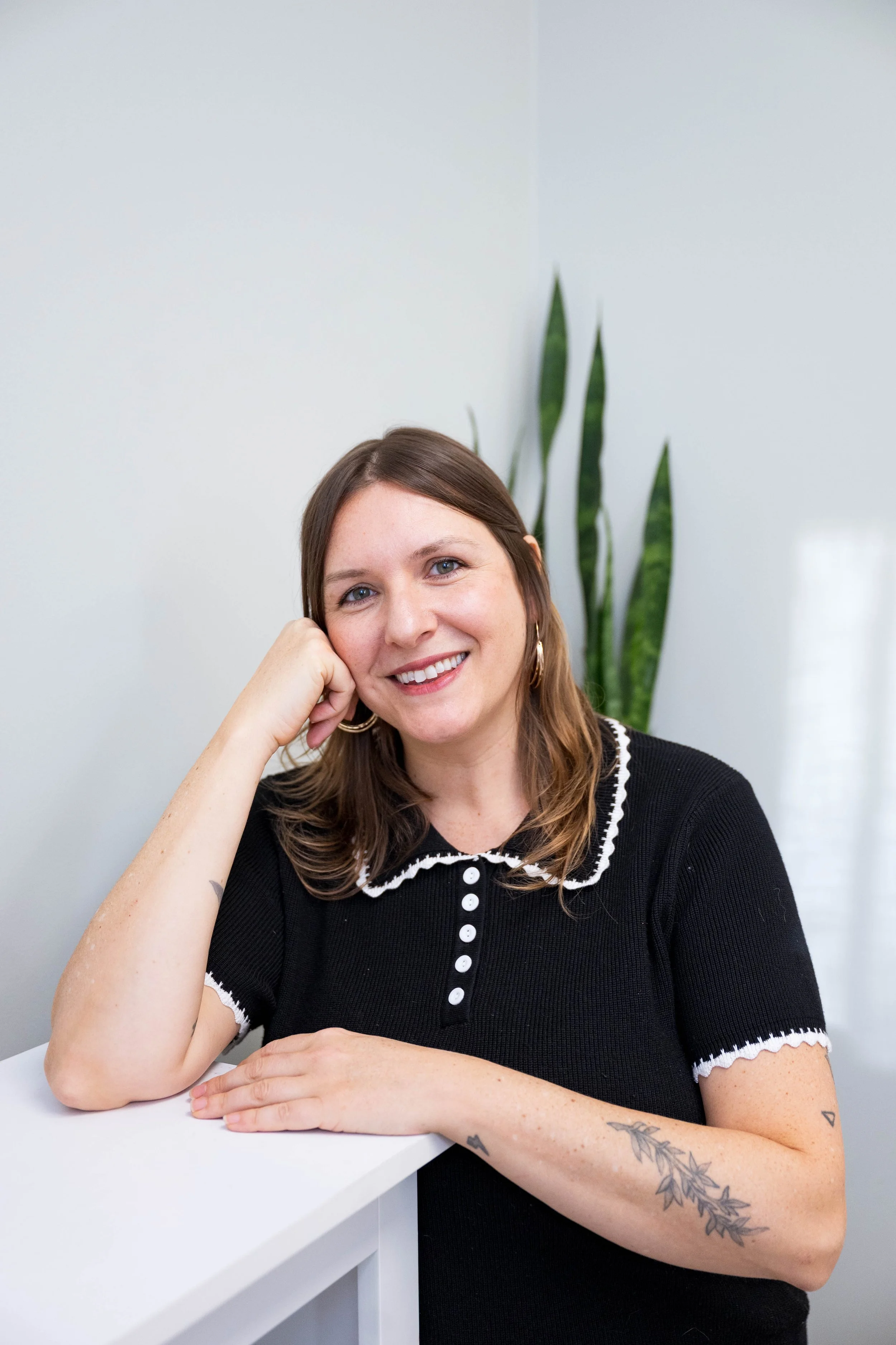 A woman with shoulder-length brown hair and tattoos on her arm, smiling and resting her head on her hand, sitting at a white table with a green plant in the background.