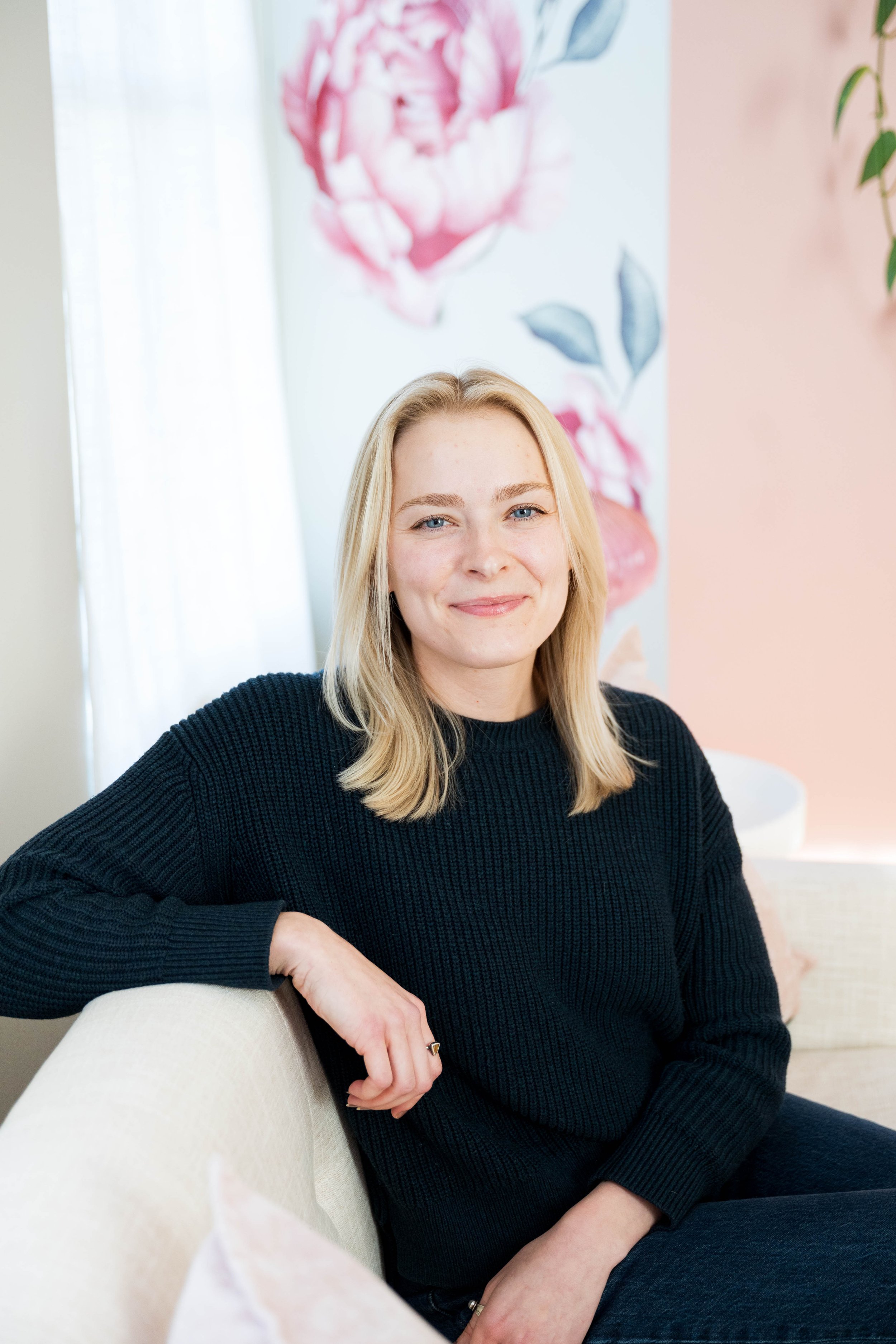 A woman with blonde hair and blue eyes wearing a black sweater, sitting on a beige couch, smiling in a bright room with floral artwork in the background.