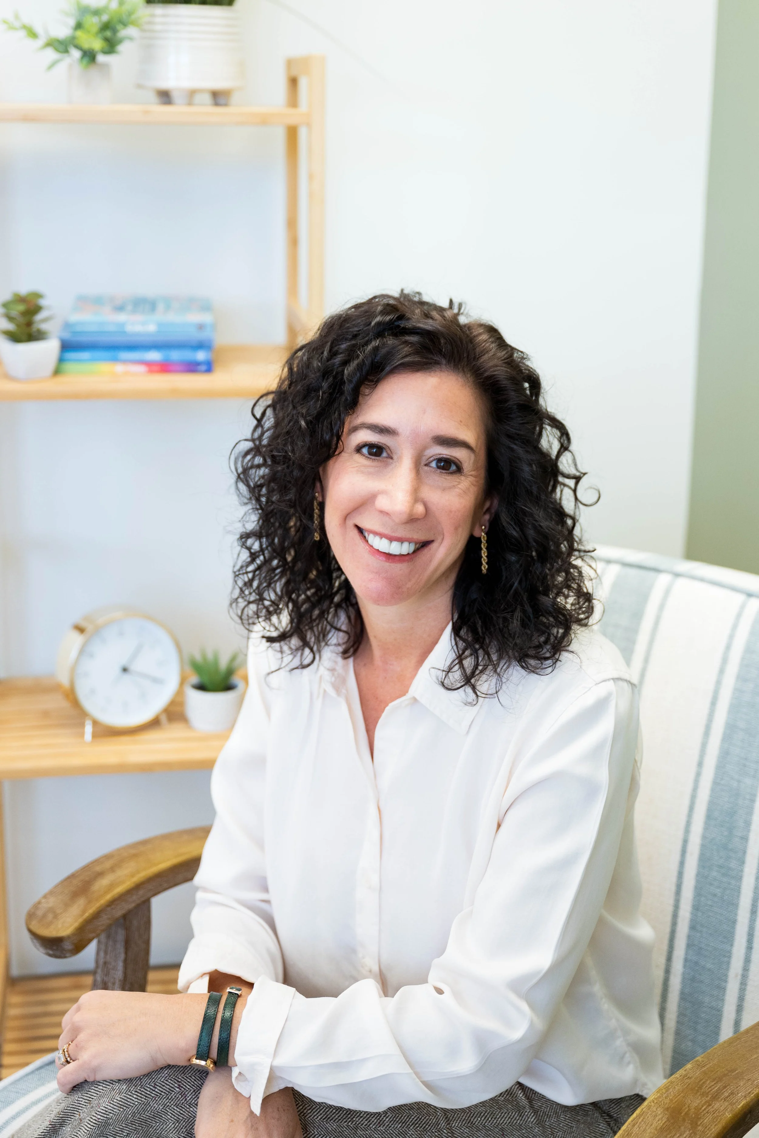 A woman with curly brown hair sitting on a gray couch, smiling at the camera. She is wearing a beige jacket and blue jeans, with artwork of leaves on the wall behind her.