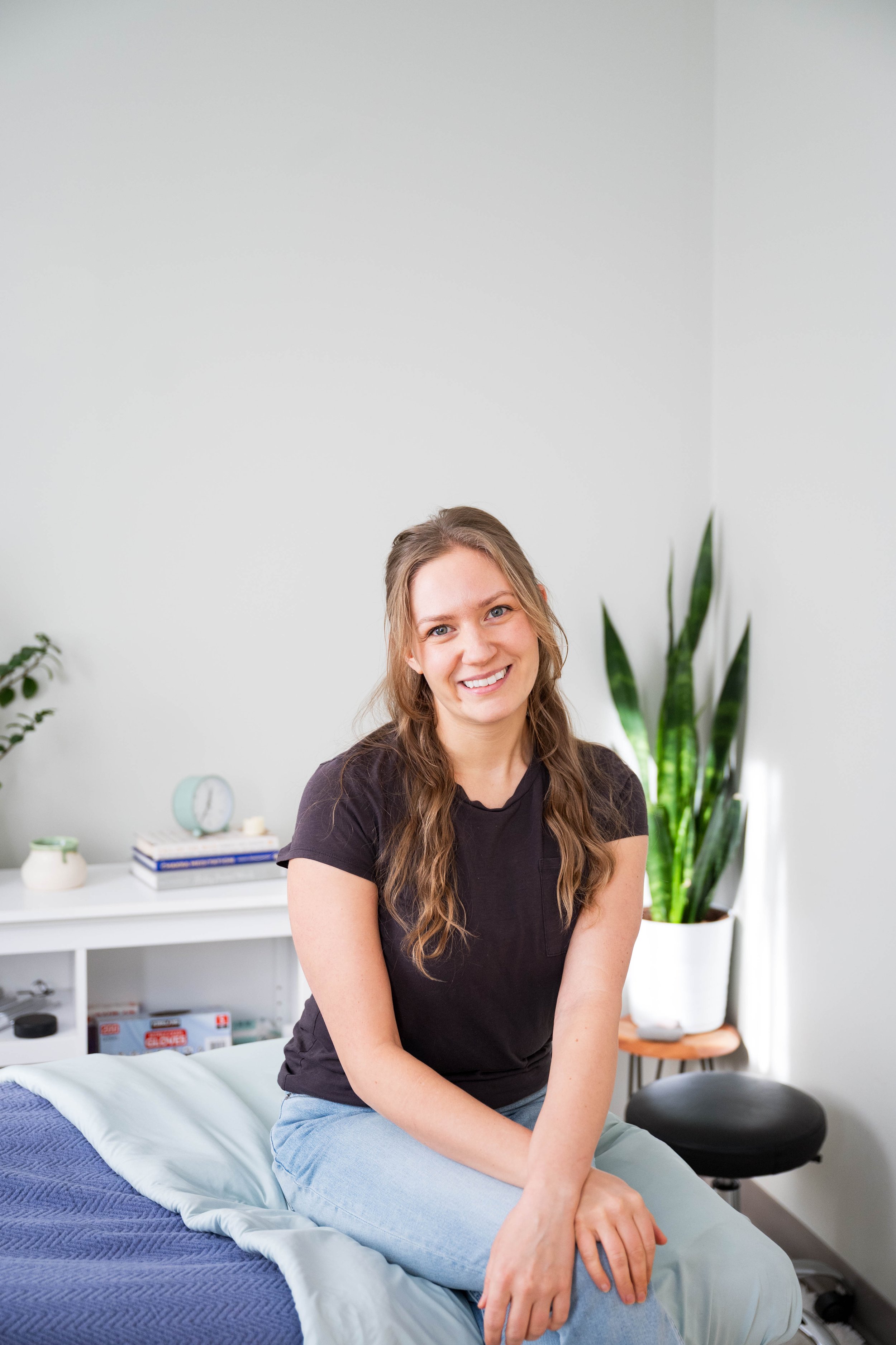 Black and white portrait of a smiling woman with wavy hair, wearing a patterned top and a cardigan, standing outdoors in front of pine branches.