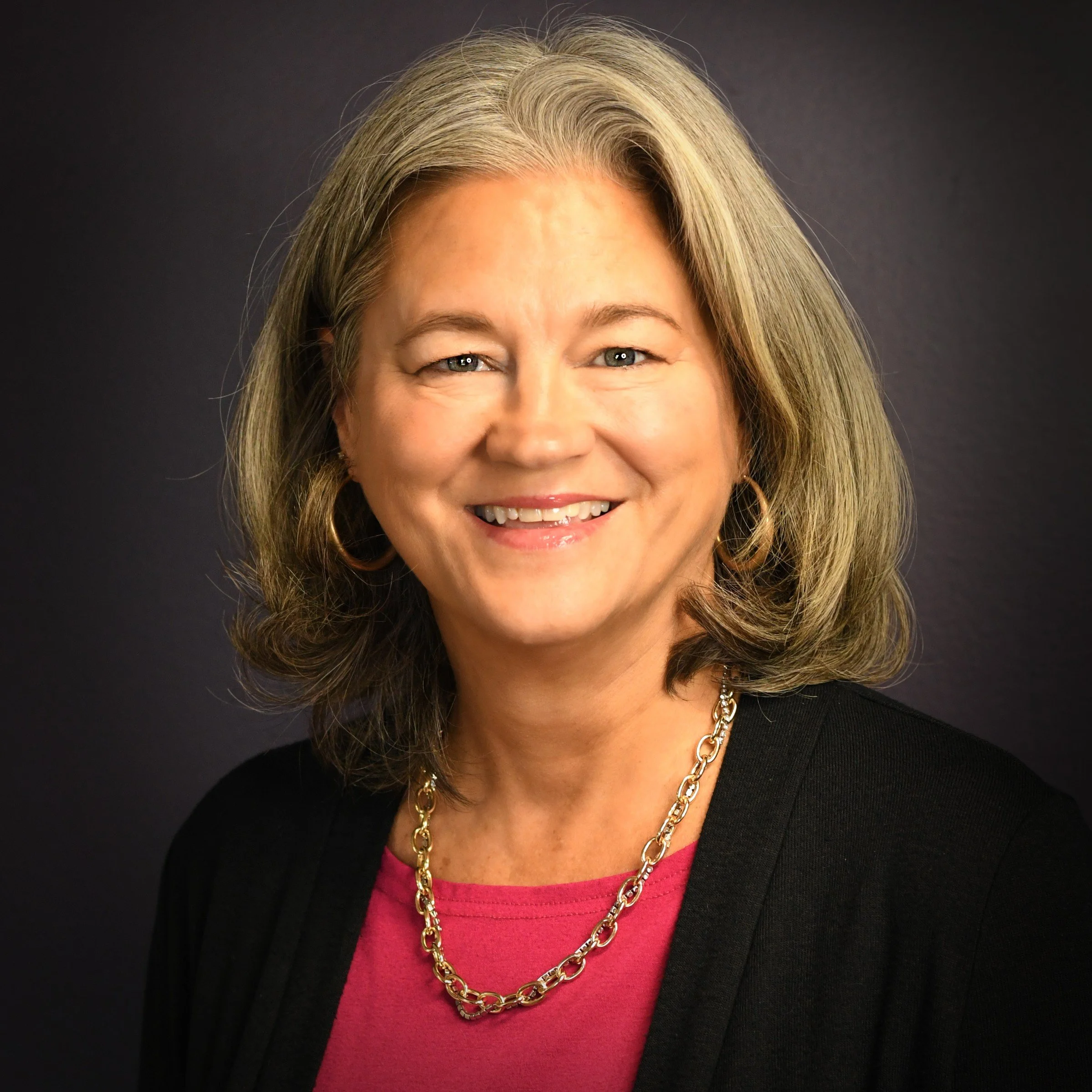 A woman with shoulder-length gray hair smiling at the camera, wearing a black shirt and a red cardigan, against a dark background.