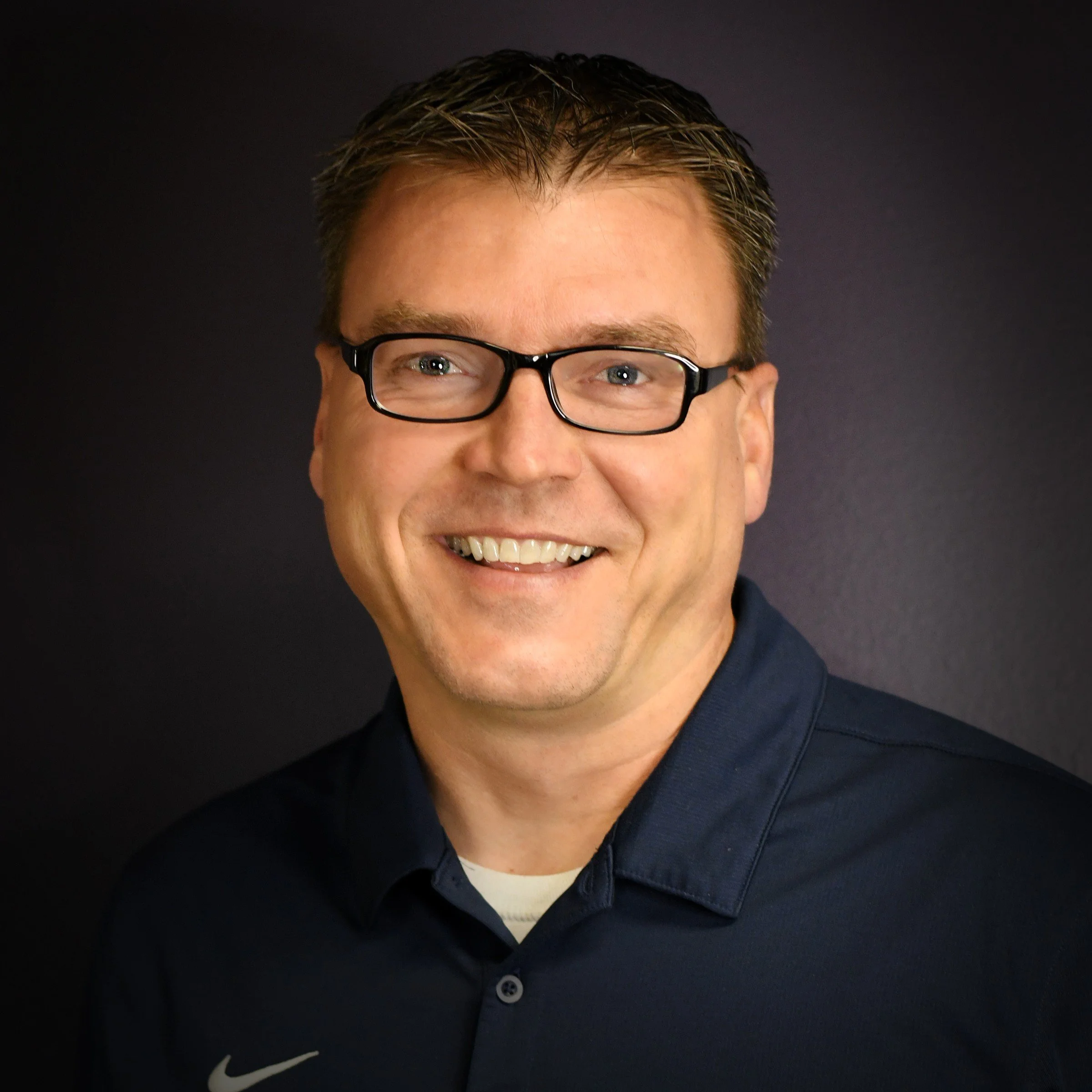 A man with brown hair, glasses, and a light gray checkered shirt standing against a dark background.