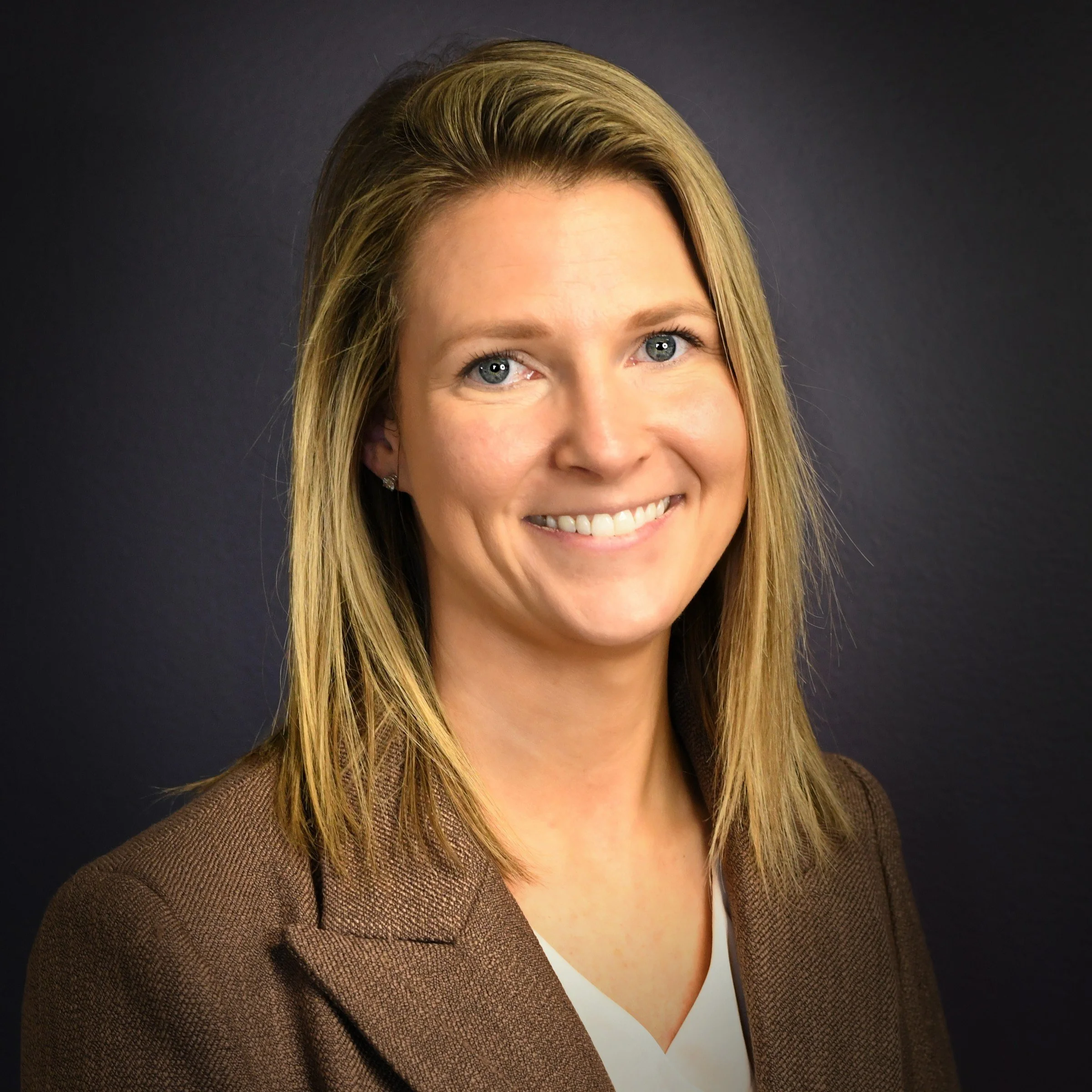 Headshot of a woman with shoulder-length blonde hair, smiling, wearing a blazer against a dark background.