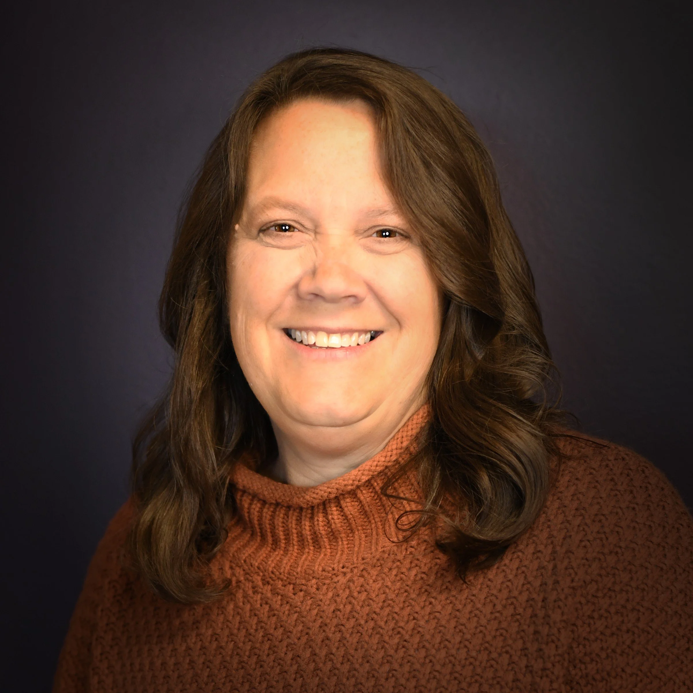 A woman with shoulder-length brown hair, wearing a dark blazer and a light green collared shirt, smiling against a gray background.