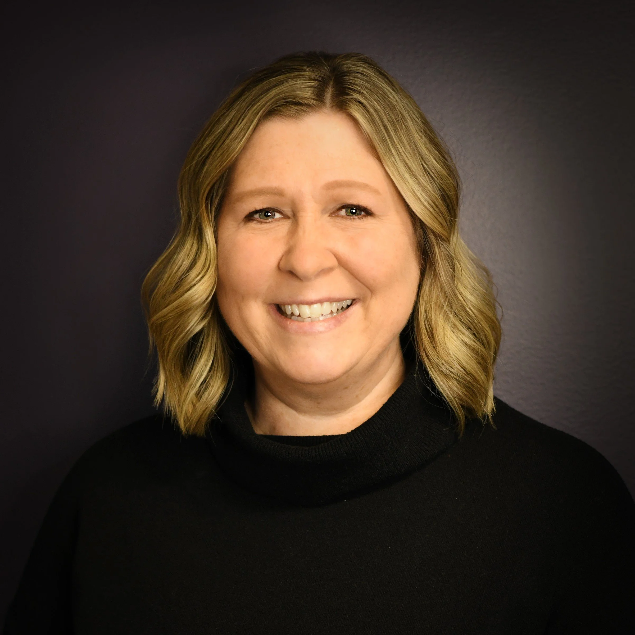 Headshot of a woman with shoulder-length blonde hair, glasses, smiling, wearing a black top, against a dark background.