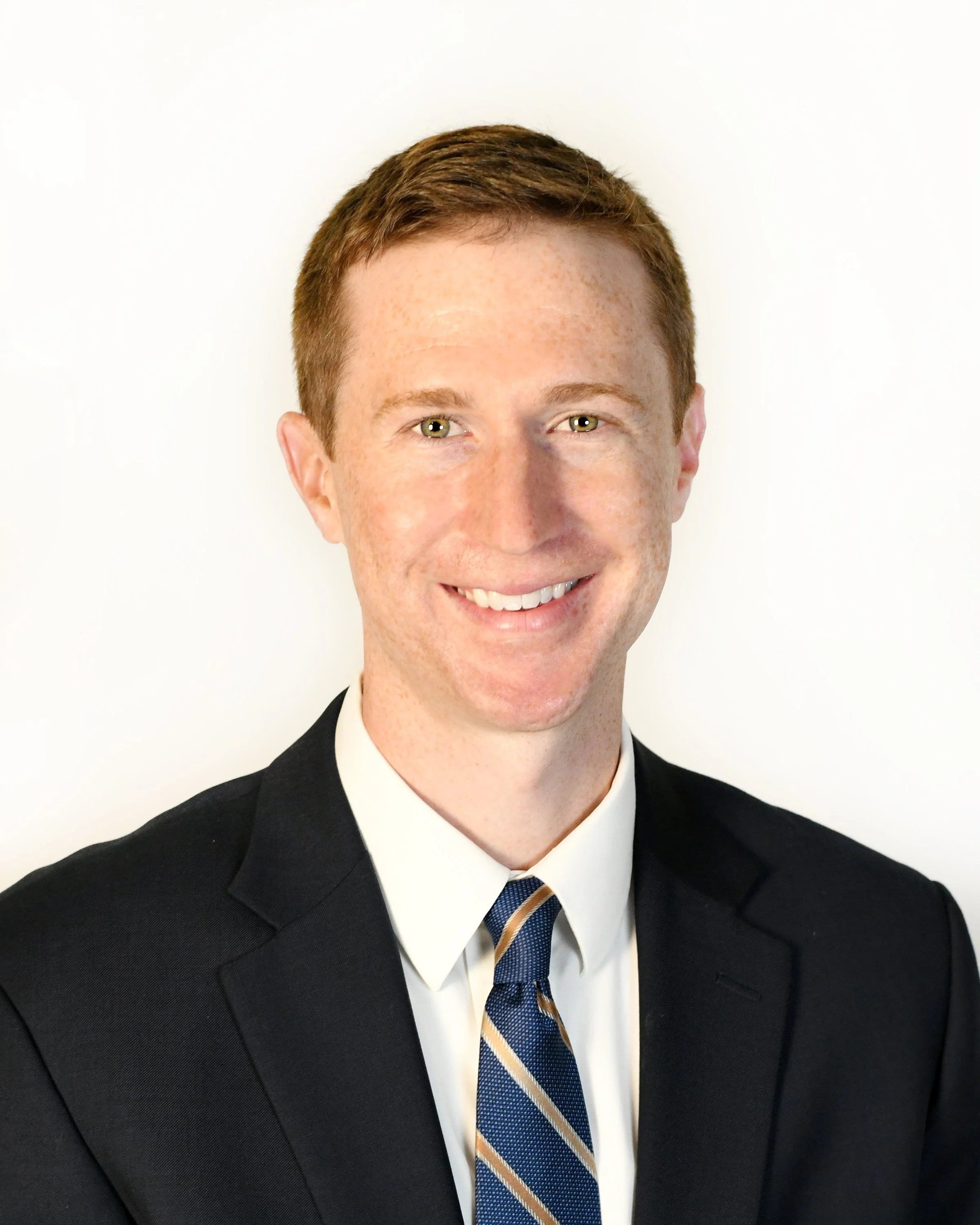 A close-up professional headshot of a smiling red-haired man wearing a suit and tie, against a plain white background.