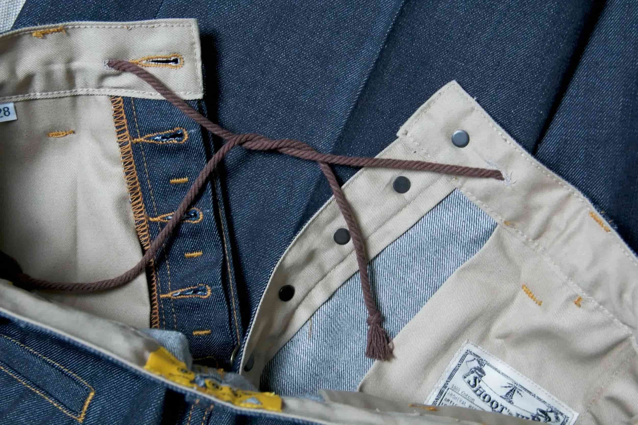 Close-up of the interior waistband of a pair of blue denim jeans showing the button, zipper, and beige inner lining with a label, along with a brown drawstring.