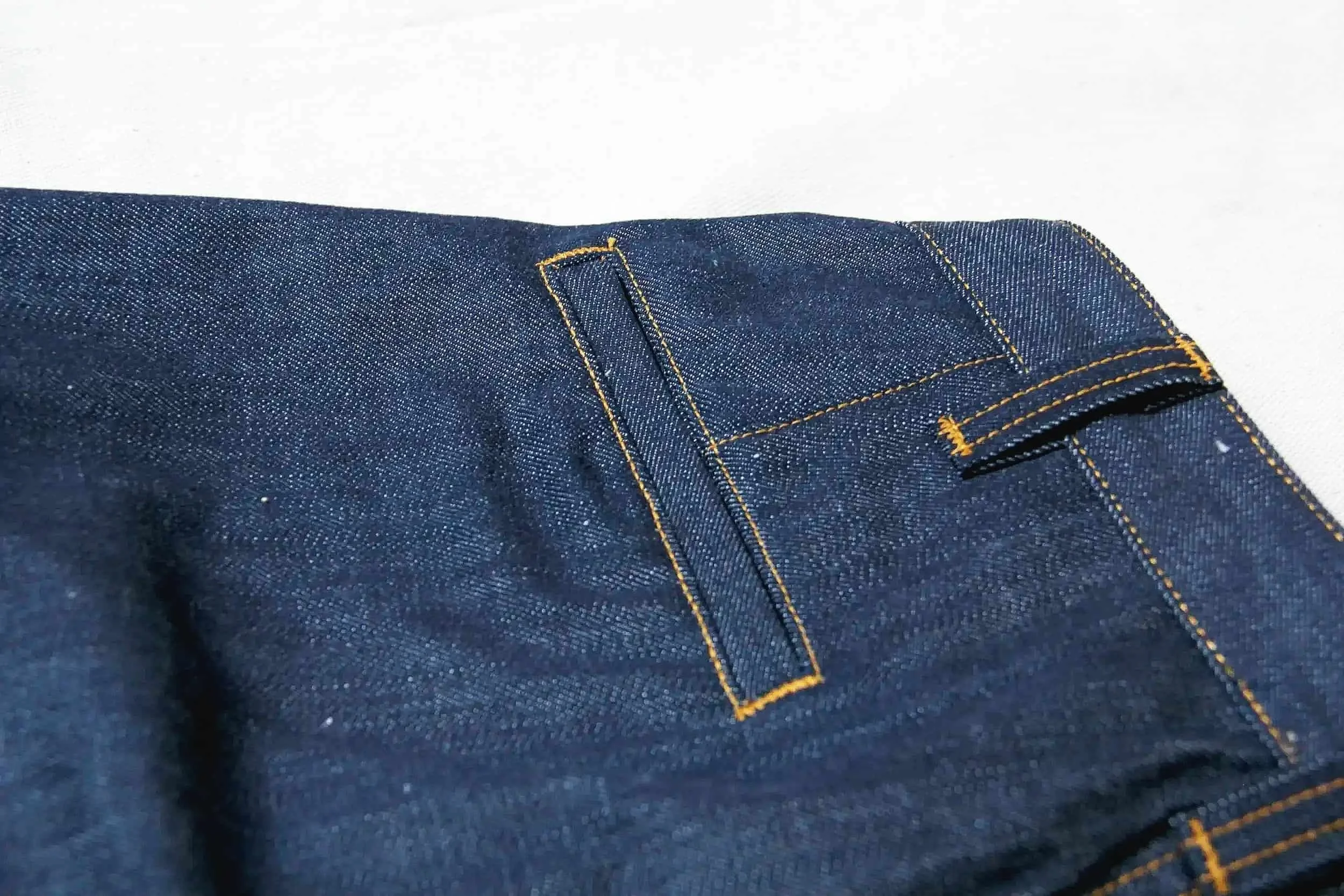 Close-up image of dark blue denim jeans with orange stitching, showing part of a back pocket.