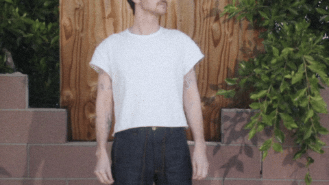 Person with short hair and tattoos wearing a white cropped t-shirt and dark jeans standing in front of a wooden background with greenery.