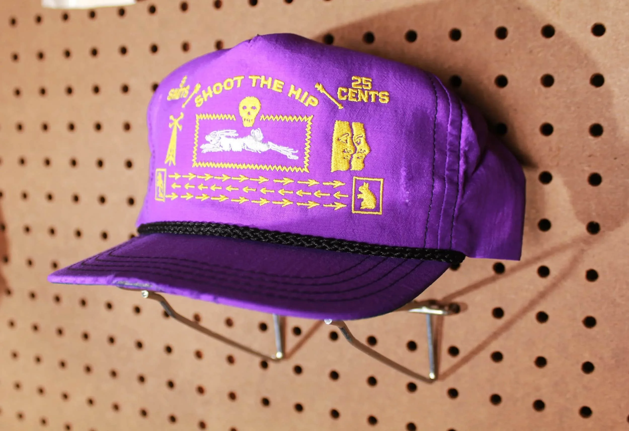 A purple cap with yellow embroidery displaying a design inspired by the Grateful Dead, featuring a skeleton lying down, text 'Shoot the Hip,' and various symbols, placed on a pegboard background.
