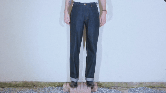 Person wearing cuffed dark blue jeans, standing on a wooden platform against a plain wall.