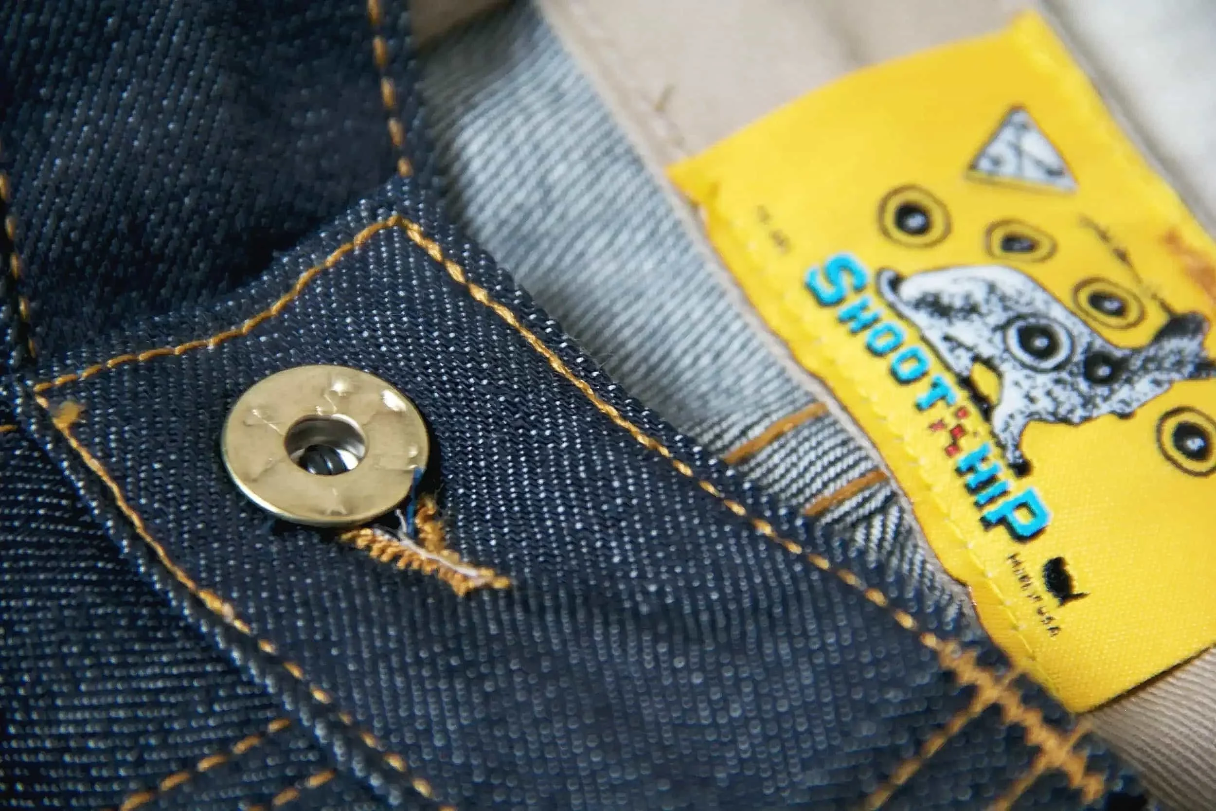 Close-up of denim fabric with yellow stitching and a metal button, alongside a yellow tag from the brand 'Snoop Dogg, Hip' featuring a cartoon dog's face with multiple eyes.