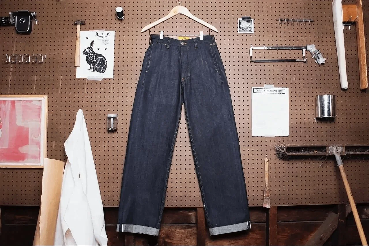 Pair of dark blue jeans hanging on a wooden hanger in a workshop with tools and materials on a pegboard wall.