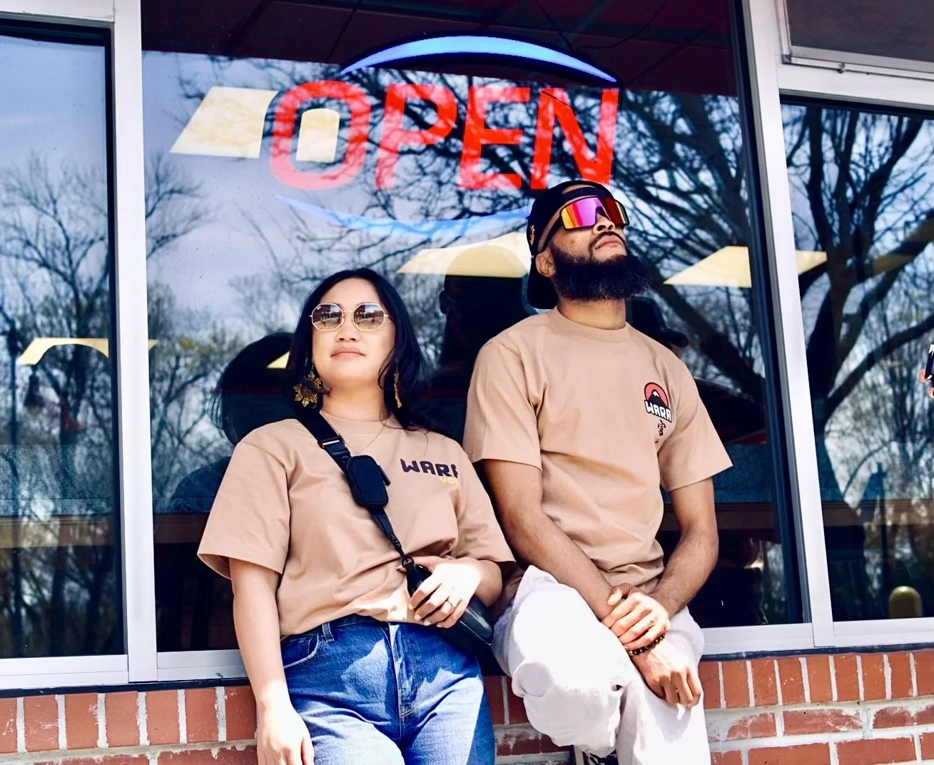 Two people standing in front of a store window with an illuminated 'OPEN' sign. The woman on the left has long dark hair, sunglasses, and a beige shirt, holding a camera. The man on the right has a beard, sunglasses, a black cap, beige shirt, and white pants, with his hands clasped in front of him.