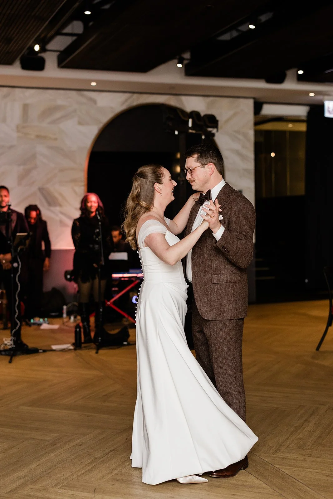 bride and a groom on a wooden dance floor smiling at each other as they perform their first dance with a live band behind them.