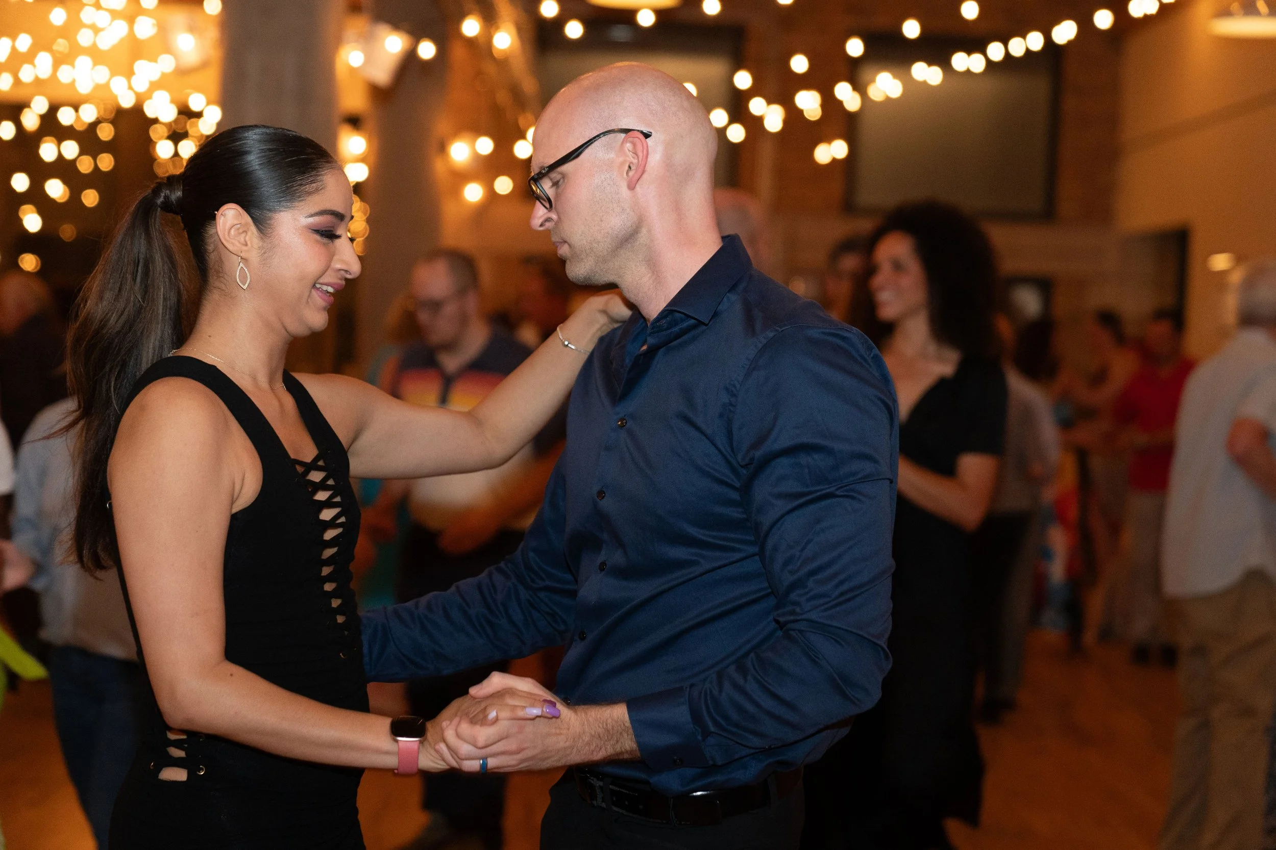 A man and woman dancing closely at a social event, with string lights in the background and other people in the room.