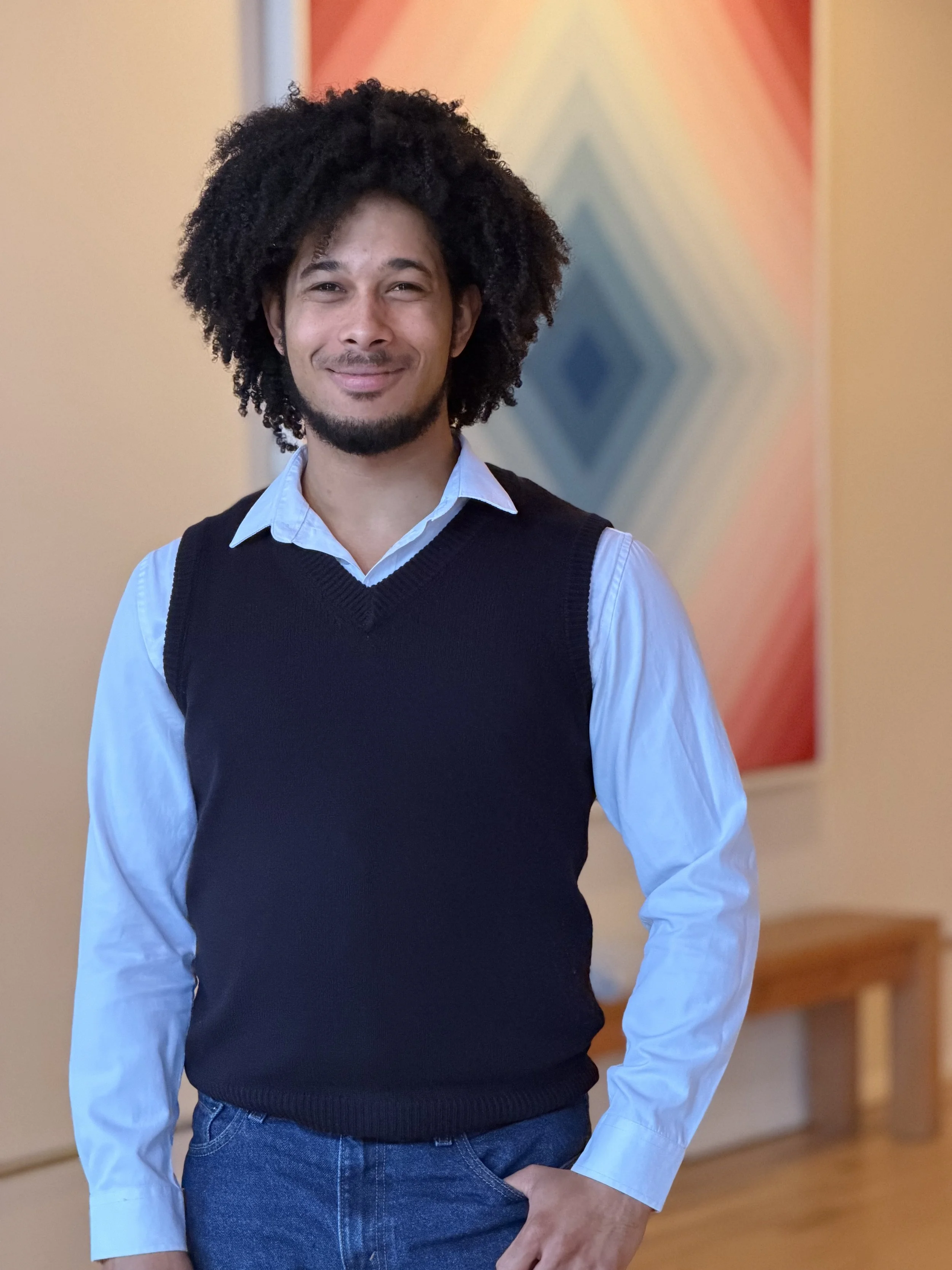 A man with curly black hair and a goatee smiling, wearing a light blue collared shirt and a black sweater vest, standing in front of an abstract geometric wall art.