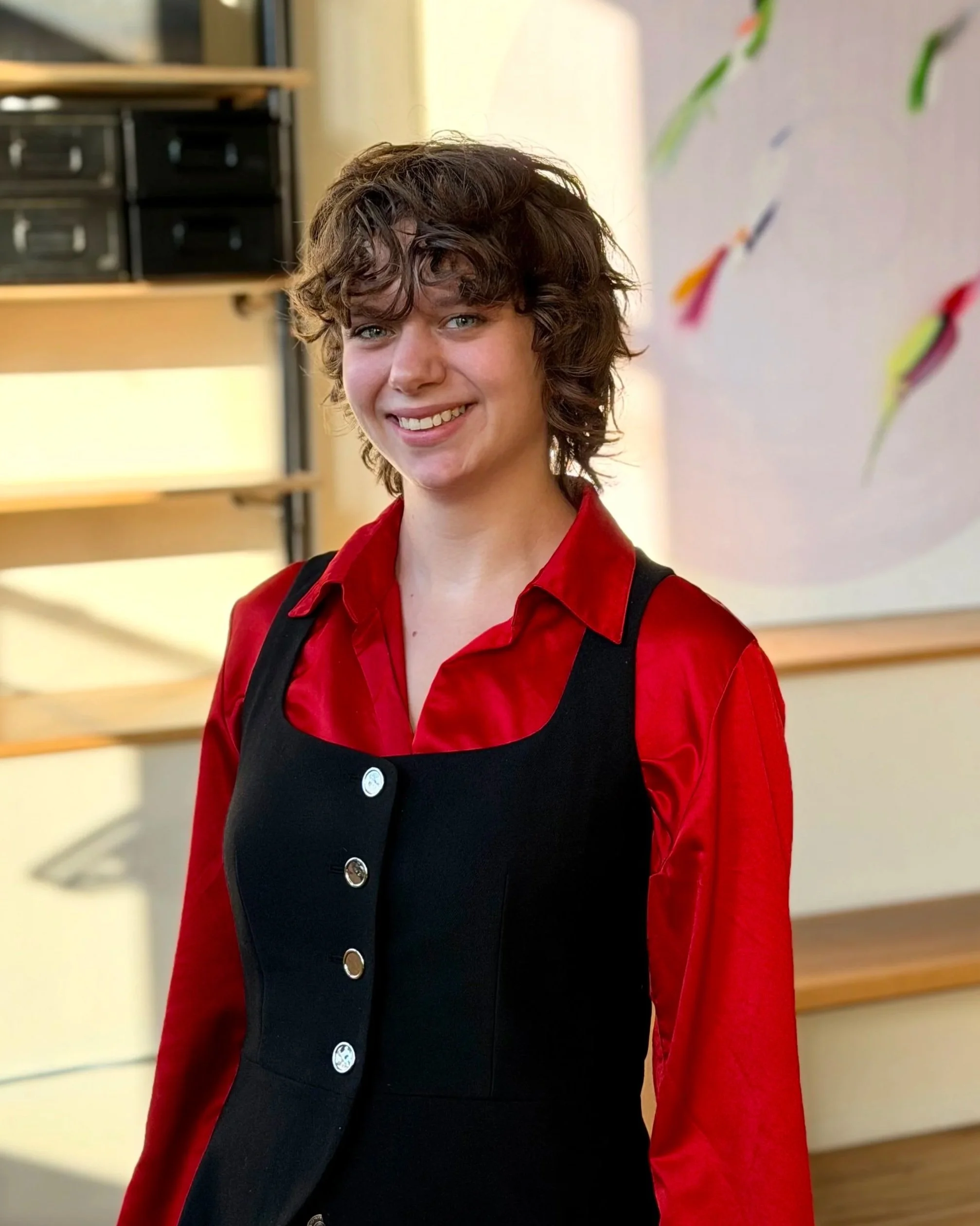 Young person with curly brown hair smiling, wearing a red shirt and a black vest, standing indoors with blurred background including a white wall with colored abstract designs.