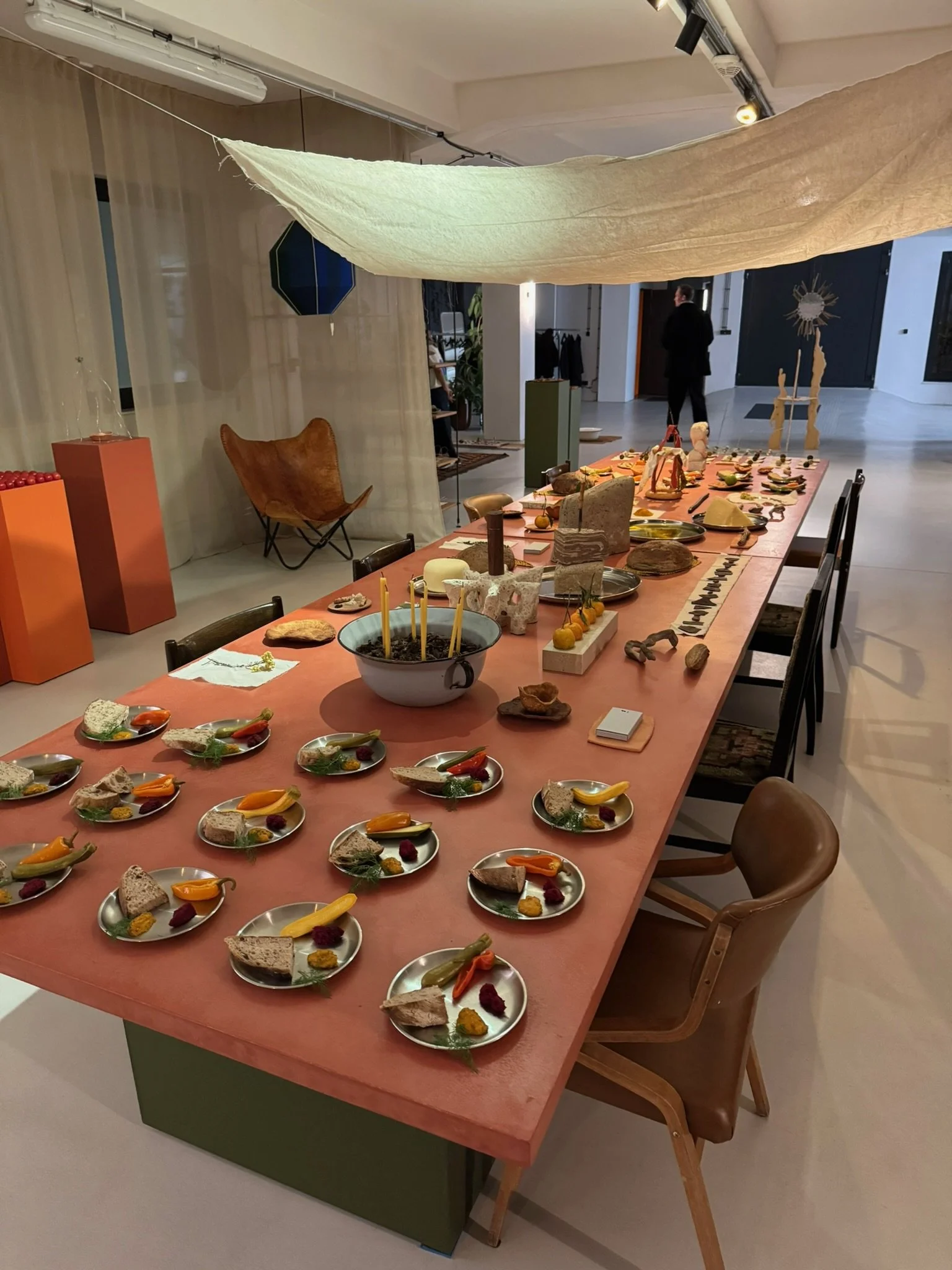 A long table with a pink tablecloth displays small plates of assorted food items, including bread, vegetables, and garnishes, in a modern art gallery or exhibition space. The background features a butterfly-shaped sculpture, a carved wooden figure, a
