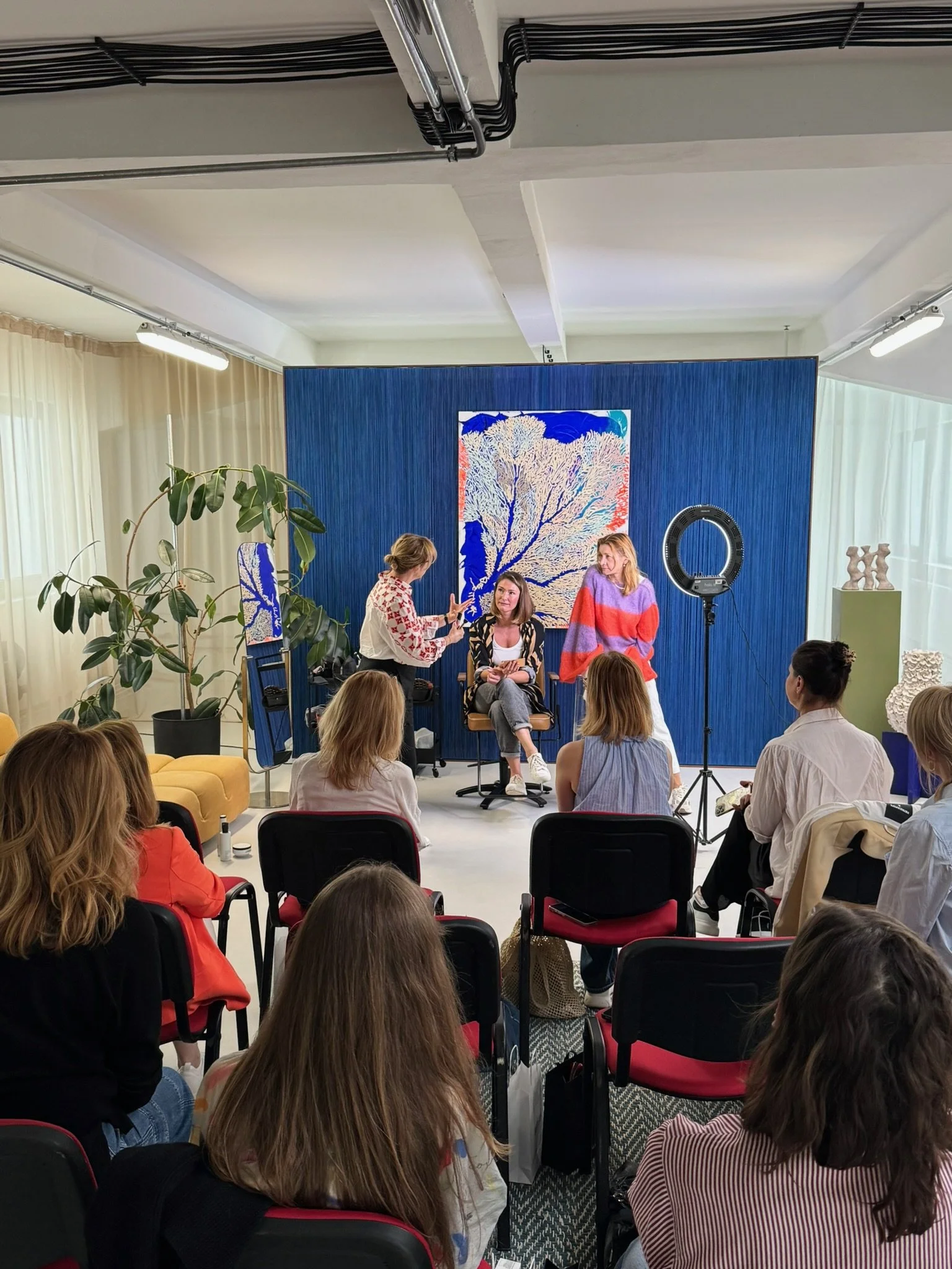 A group of women attending a workshop or presentation indoors. Three women are in the front, one sitting in a chair and two standing, engaged in conversation. The audience is seated and listening. There is a large artwork of a tree on a blue backdrop