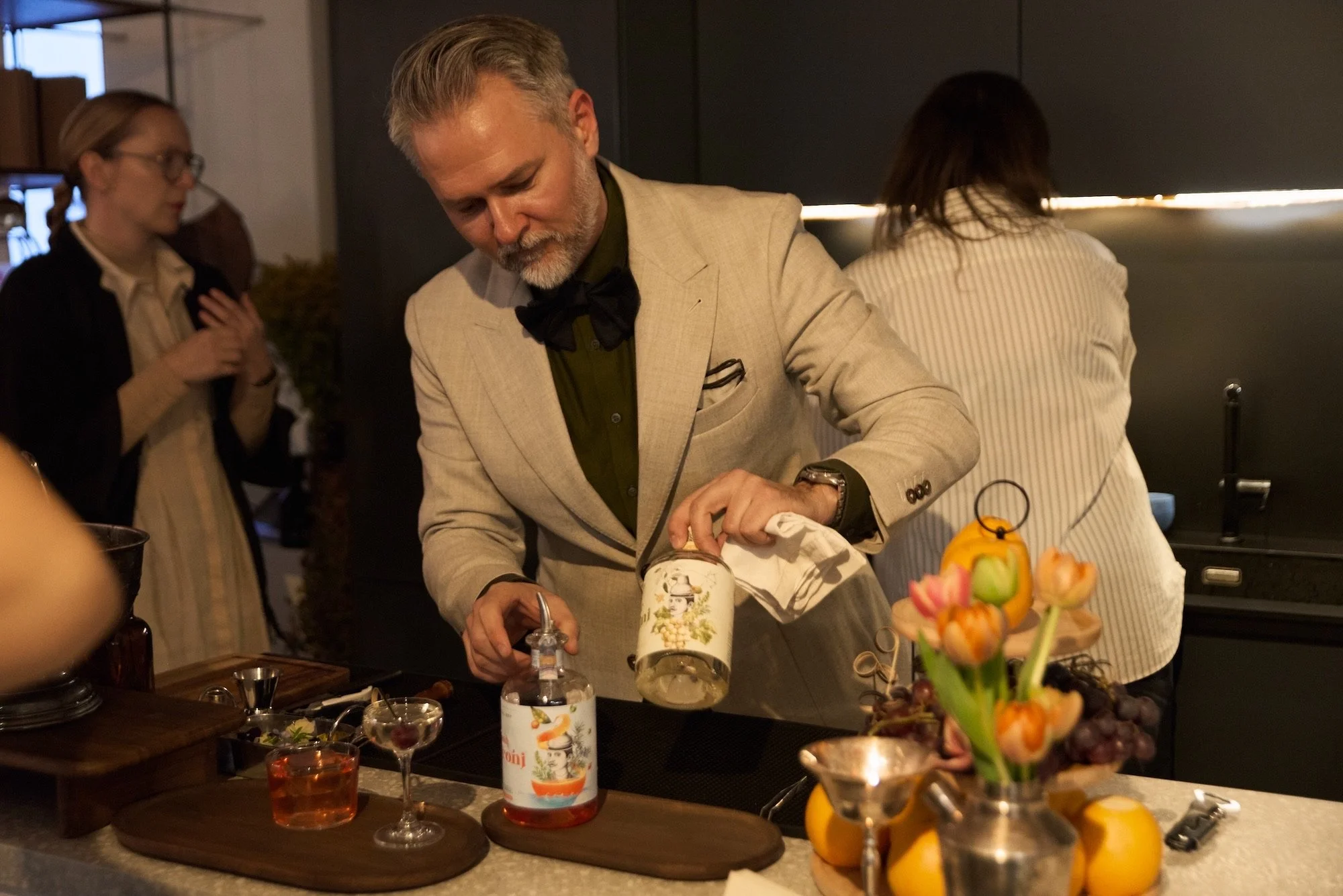 A man in a beige suit and black bow tie pouring a liquid into a small bottle at a kitchen counter during a gathering. Other people are visible in the background, and colorful tulip flowers are in a vase in the foreground.