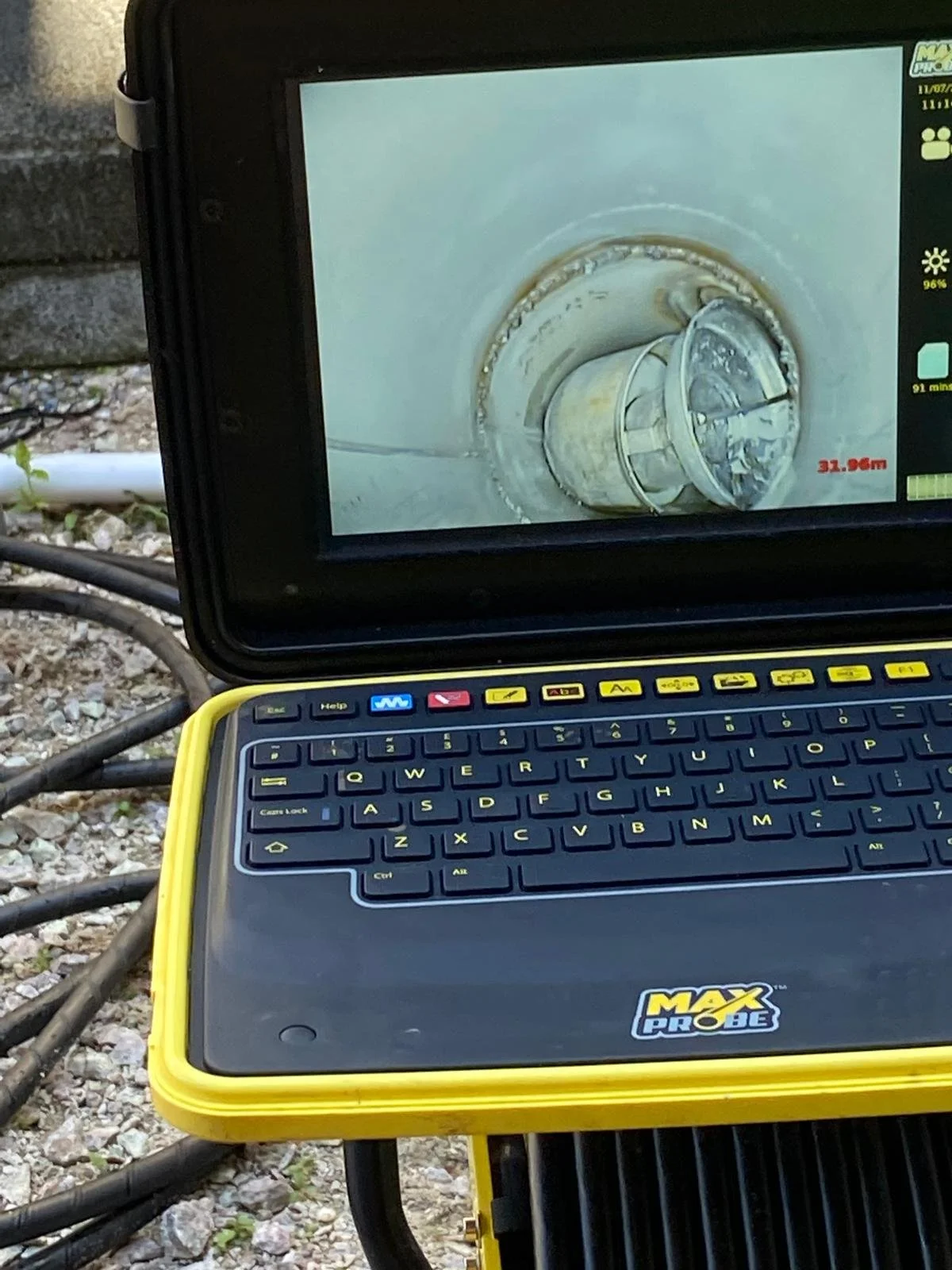 A portable sewer inspection camera with a screen displaying an interior view of a pipe. The device features a keyboard and is branded with "MAX PROBE." Cables are visible in the background on a gravel surface.
