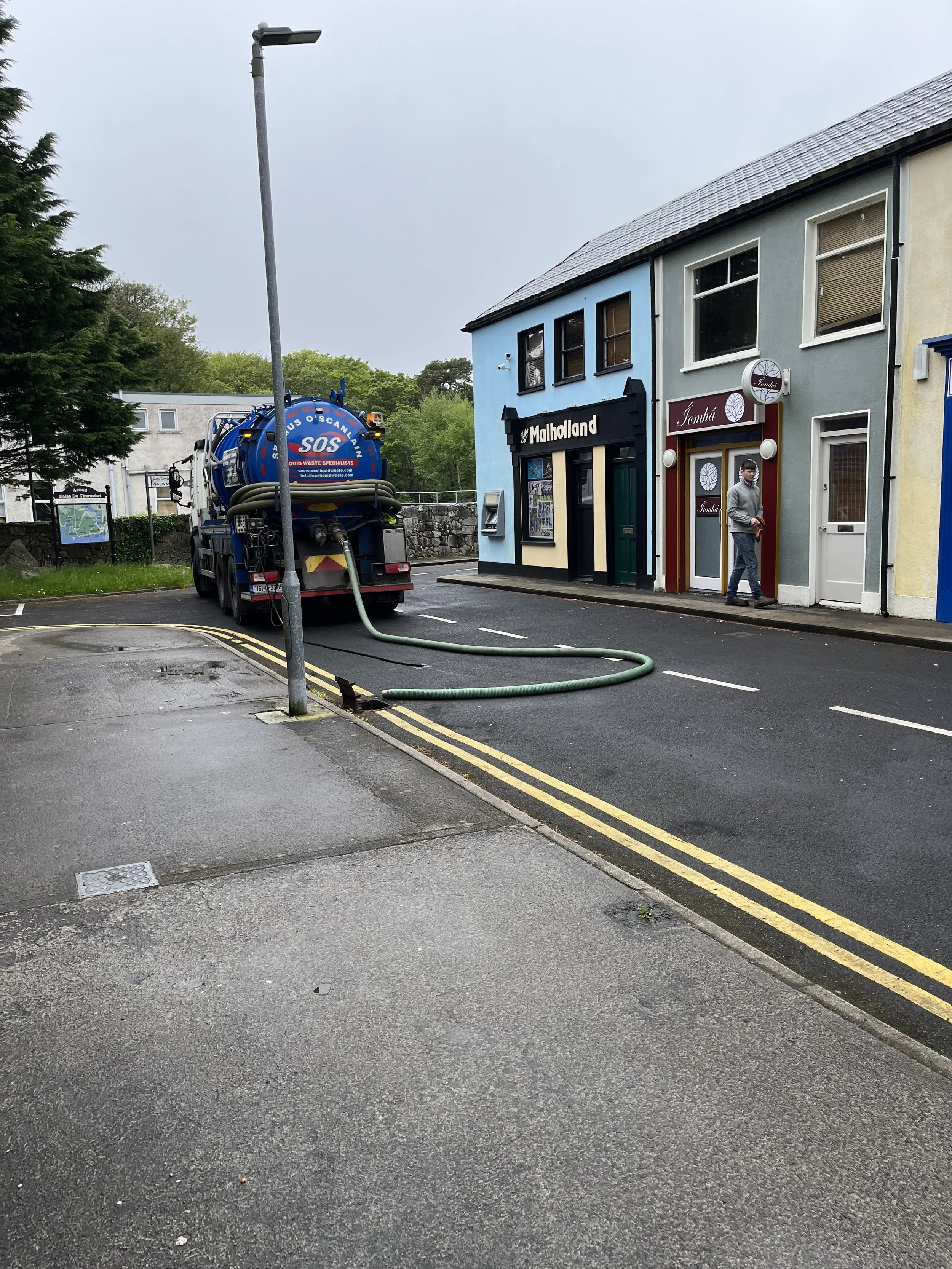 A septic tank service truck parked on a street next to buildings, with a hose extending from the truck to the building, suggesting a sewage or waste water extraction. A person is walking by on the sidewalk.