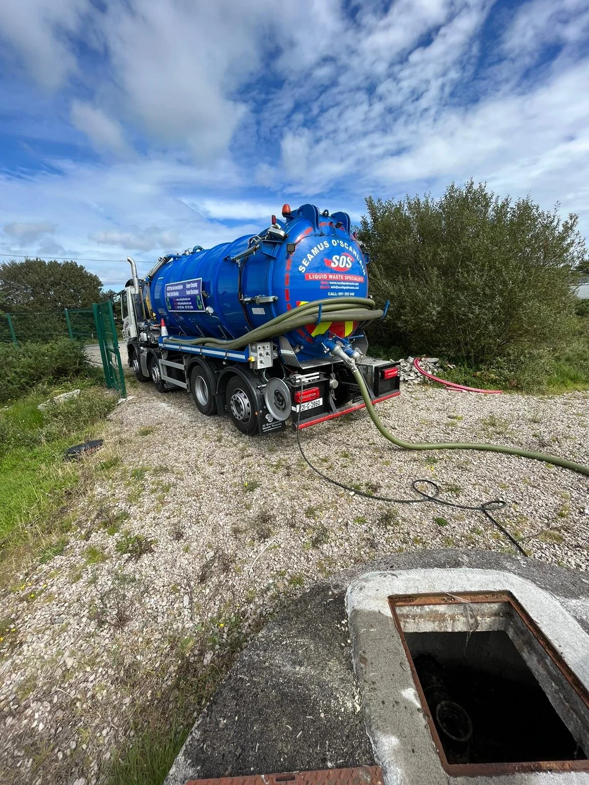 Blue liquid waste tanker truck parked near an open manhole on a gravel area with hoses attached, under a partly cloudy sky.