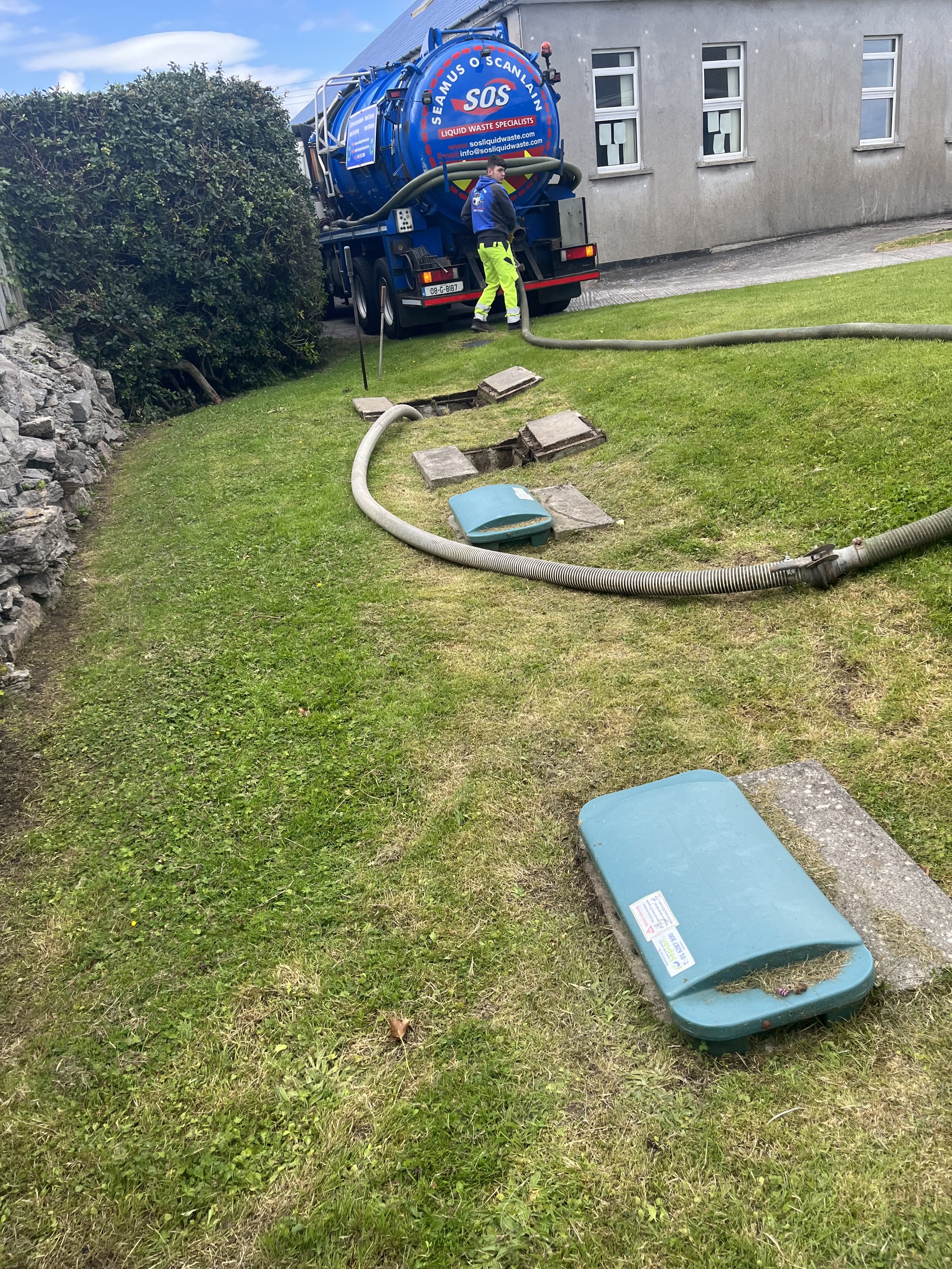 Septic tank service truck with hose attached, man in safety gear handling the hose, lawn with open septic tank covers, adjacent building.