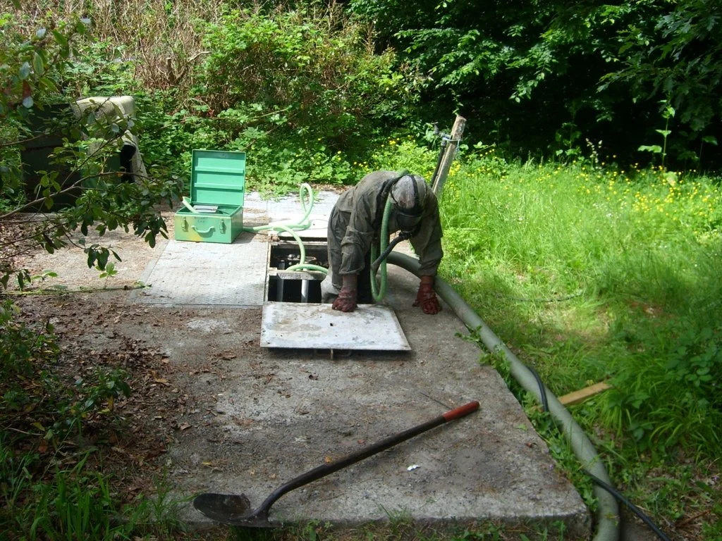Person cleaning a septic tank in a garden area, surrounded by greenery, with open hatch and tools on the ground.