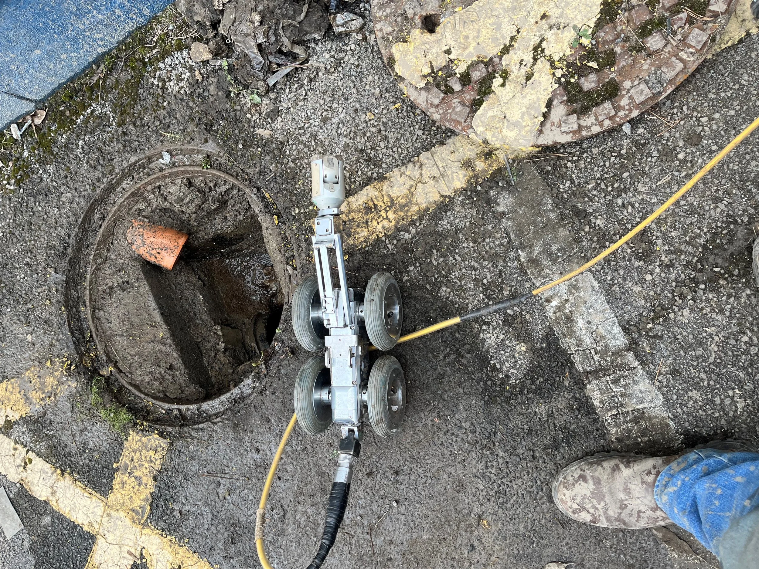 A robotic camera on wheels positioned near an open manhole, used for inspecting underground sewer lines. A cable is attached to the robot, and there is debris around the manhole edge. A person wearing jeans and muddy boots is partially visible.