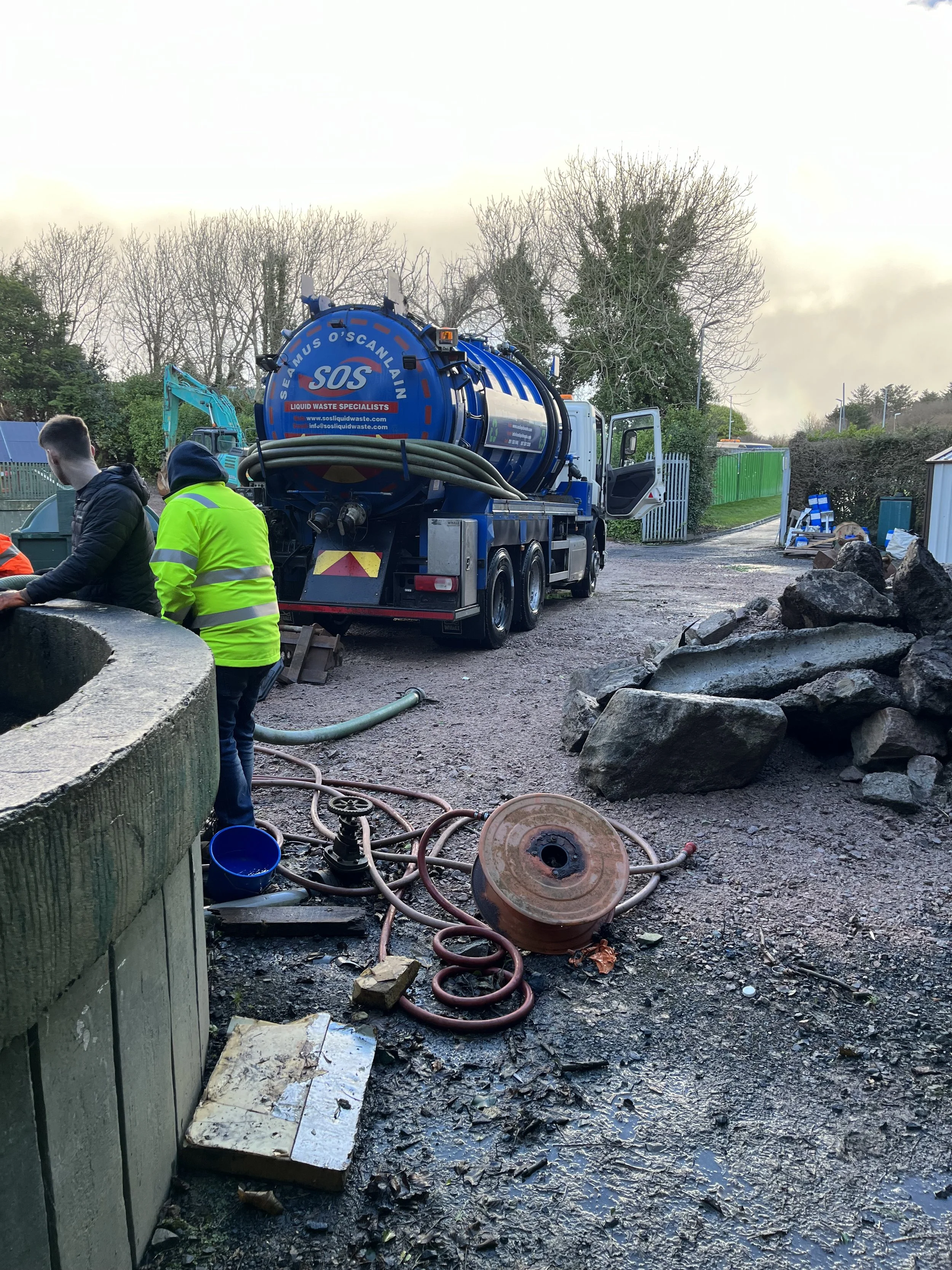 Waste management truck with hoses, workers in safety vests, and construction debris in a yard.