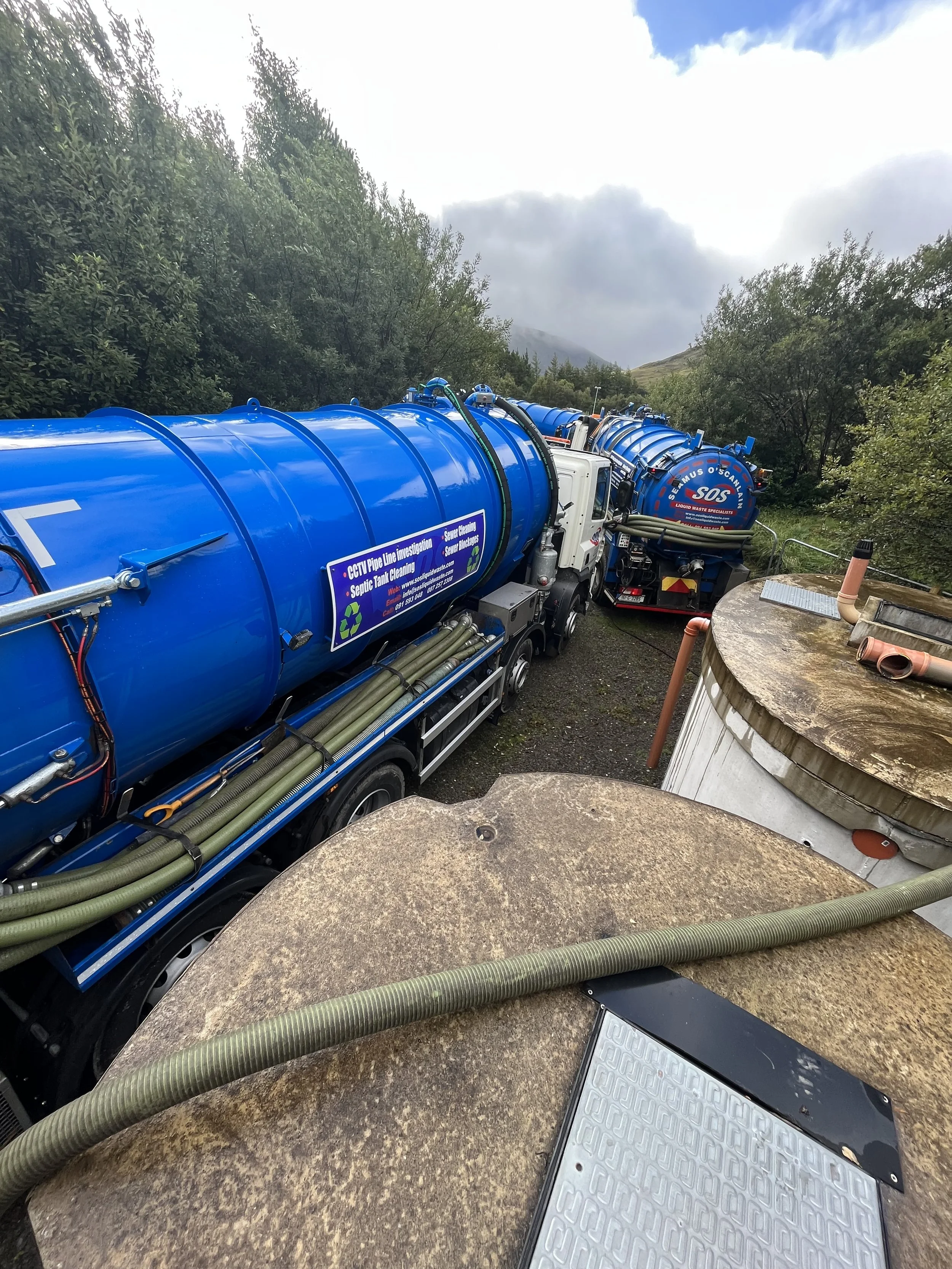 Blue SOS Liquid Waste septic tank trucks with hoses attached, parked in a wooded area with cloudy skies.