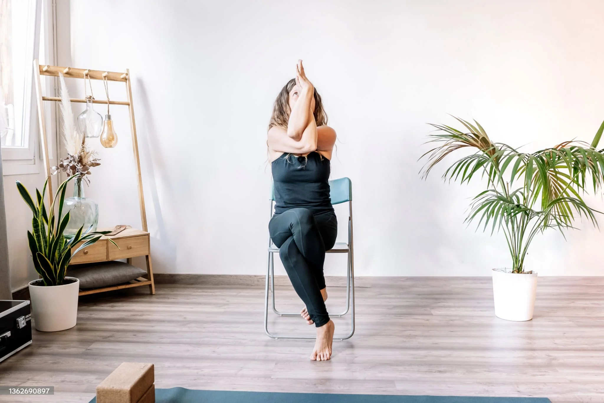 Aulas adaptadas de yoga. Mulher a praticar ioga sentada numa cadeira, a fazer a Pose da Águia, numa sala com plantas e decoração minimalista.