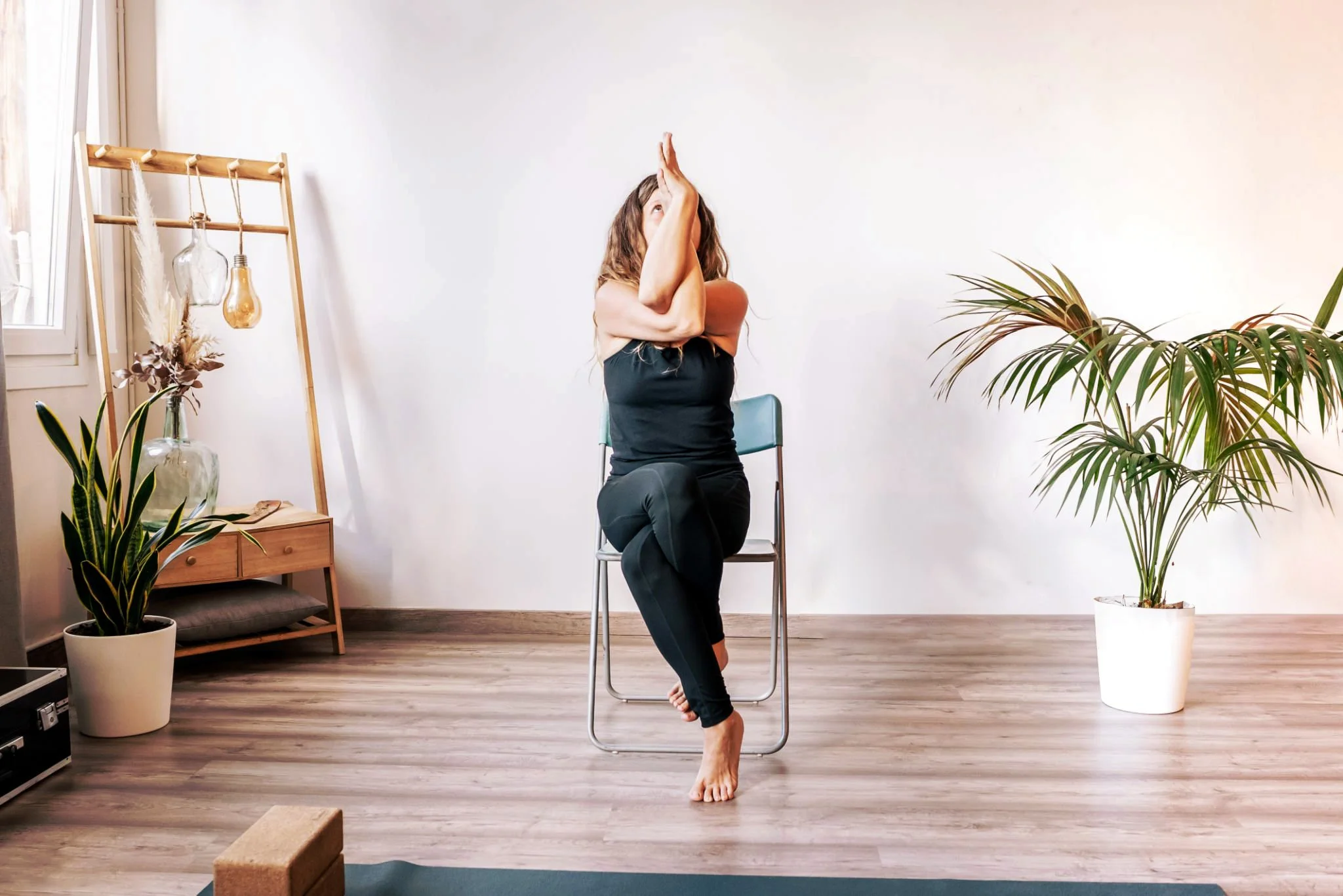 Aulas adaptadas de yoga. Mulher a praticar ioga sentada numa cadeira, a fazer a Pose da Águia, numa sala com plantas e decoração minimalista.